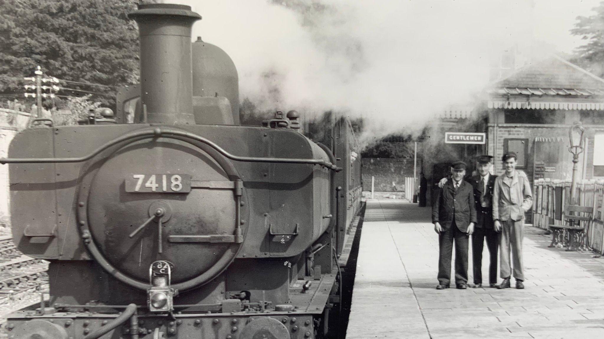 A black and white image of a steam train waiting on a platform while three men have their arms round each other smiling for a photo. They are wearing suits and hats, that look like they work at the station.