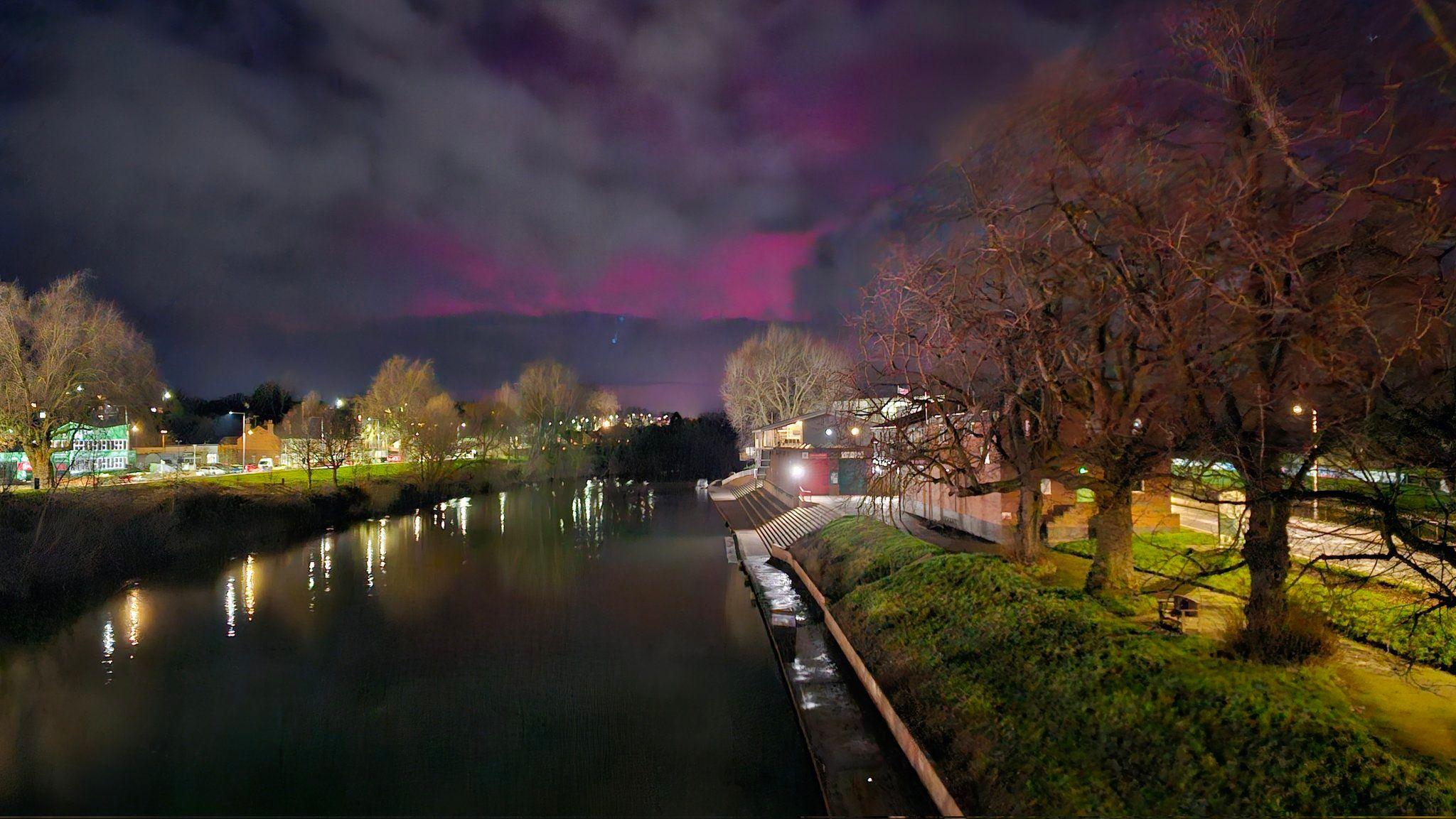 Pink hues of the aurora can be seen at the back of the photo, covered with grey clouds, over the water. Bare winter trees line both sides of the river