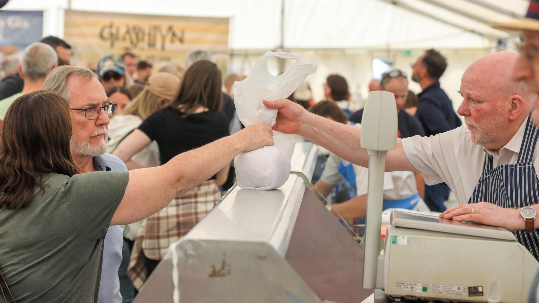 A butcher handing over goods to a couple in a marquee with crowds in the background.