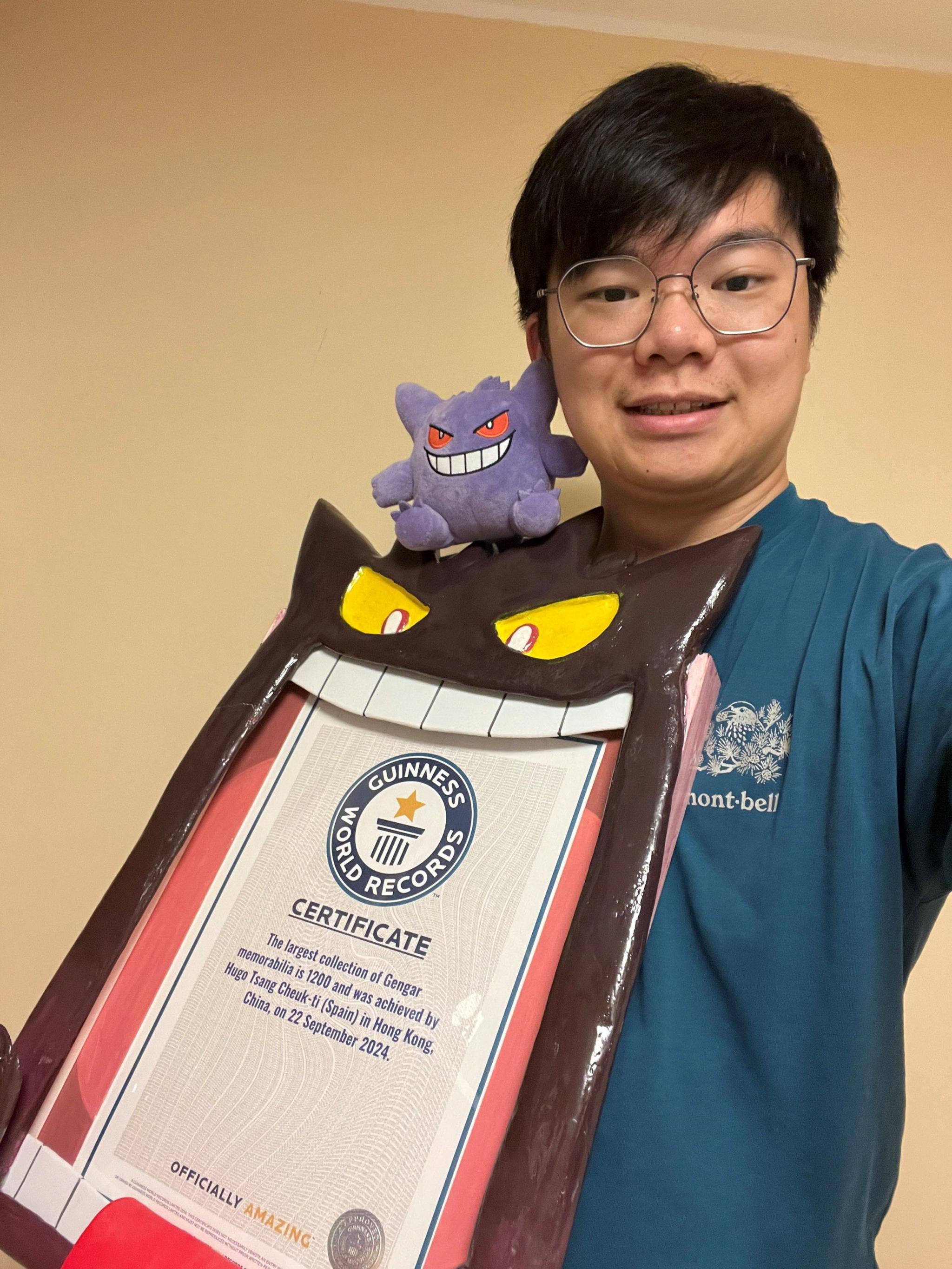 tsang smiling while holding his guinness world record certificate in a gengar frame with a plush gengar sat on his shoulder.