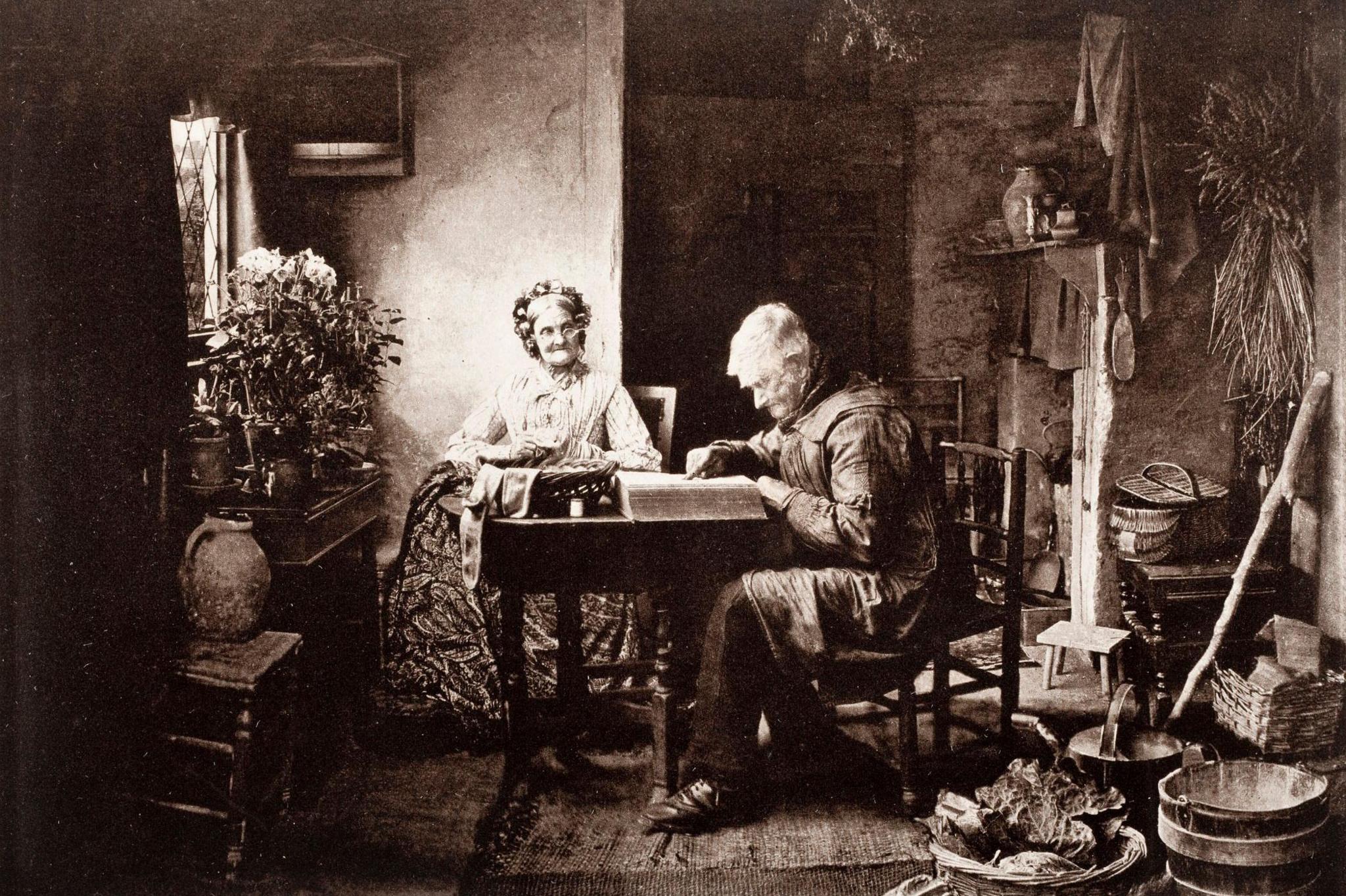 An elderly man sits in a parlour reading a book while a female figure looks on. She looks to be mending some socks. The pair are surrounded by objects relating to rural life in Victorian times, including a stool, kitchen equipment, buckets and baskets