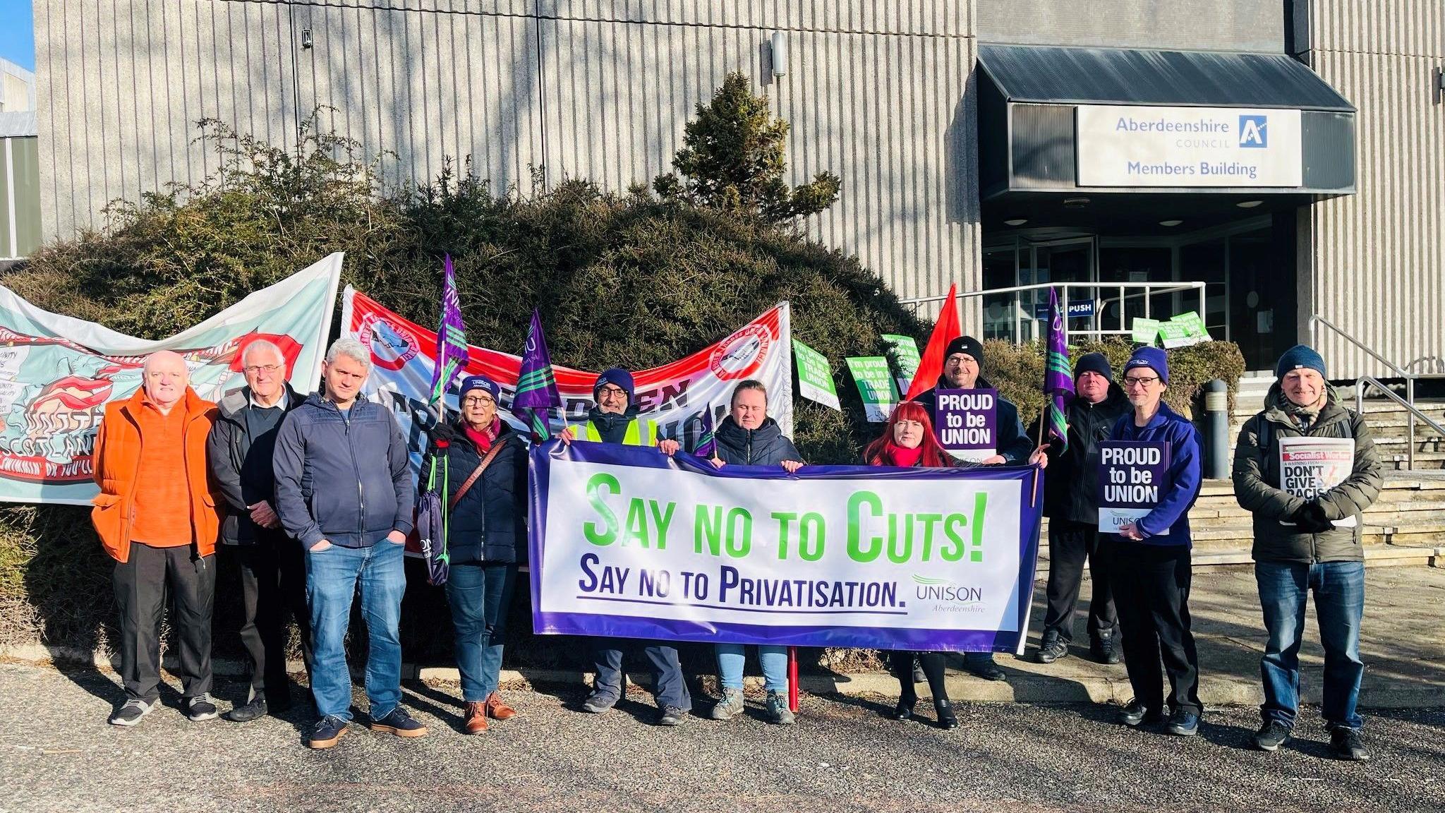 A group of people with a sign saying "Say No to Cuts!" stand outside Aberdeenshire Council on a sunny day