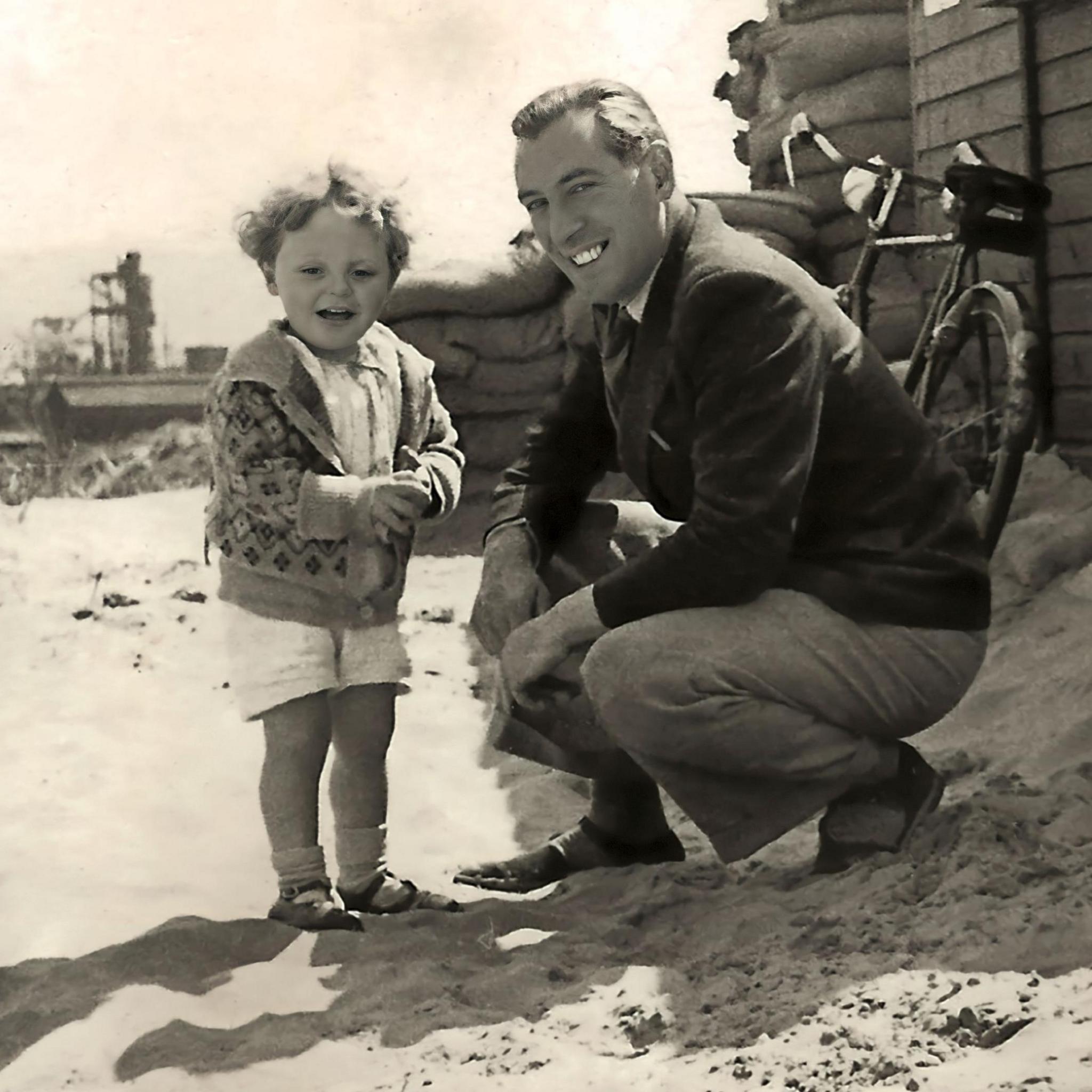 Anthony at 3, smiling, in white shorts and embroidered cardigan, curly hair, and 
his father, Richard, crouching in beige trousers and dark jacket
at Aberavon Beach in 1941 