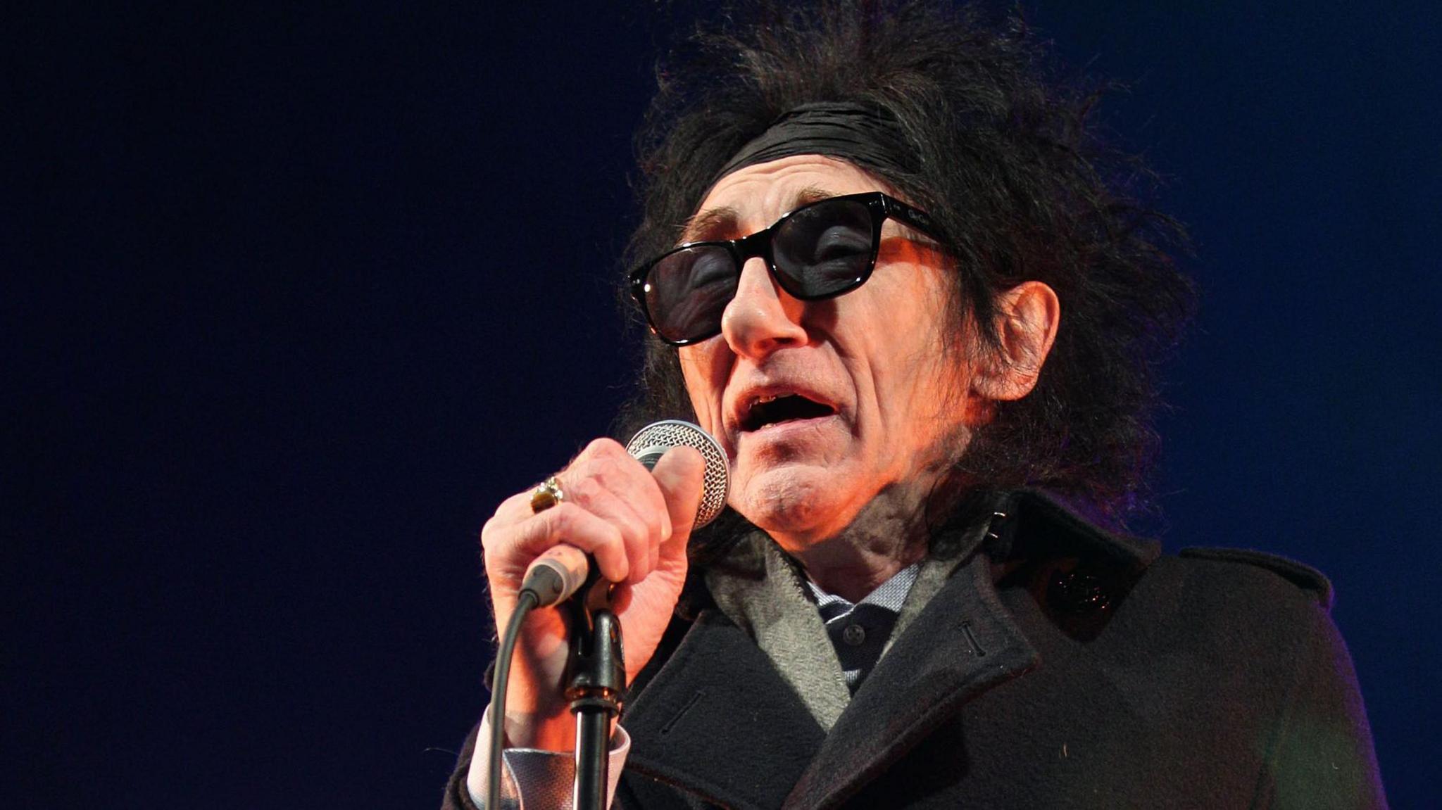 John Cooper Clarke, with wild black hair and wearing black sunglasses and a black coat, speaks into a microphone