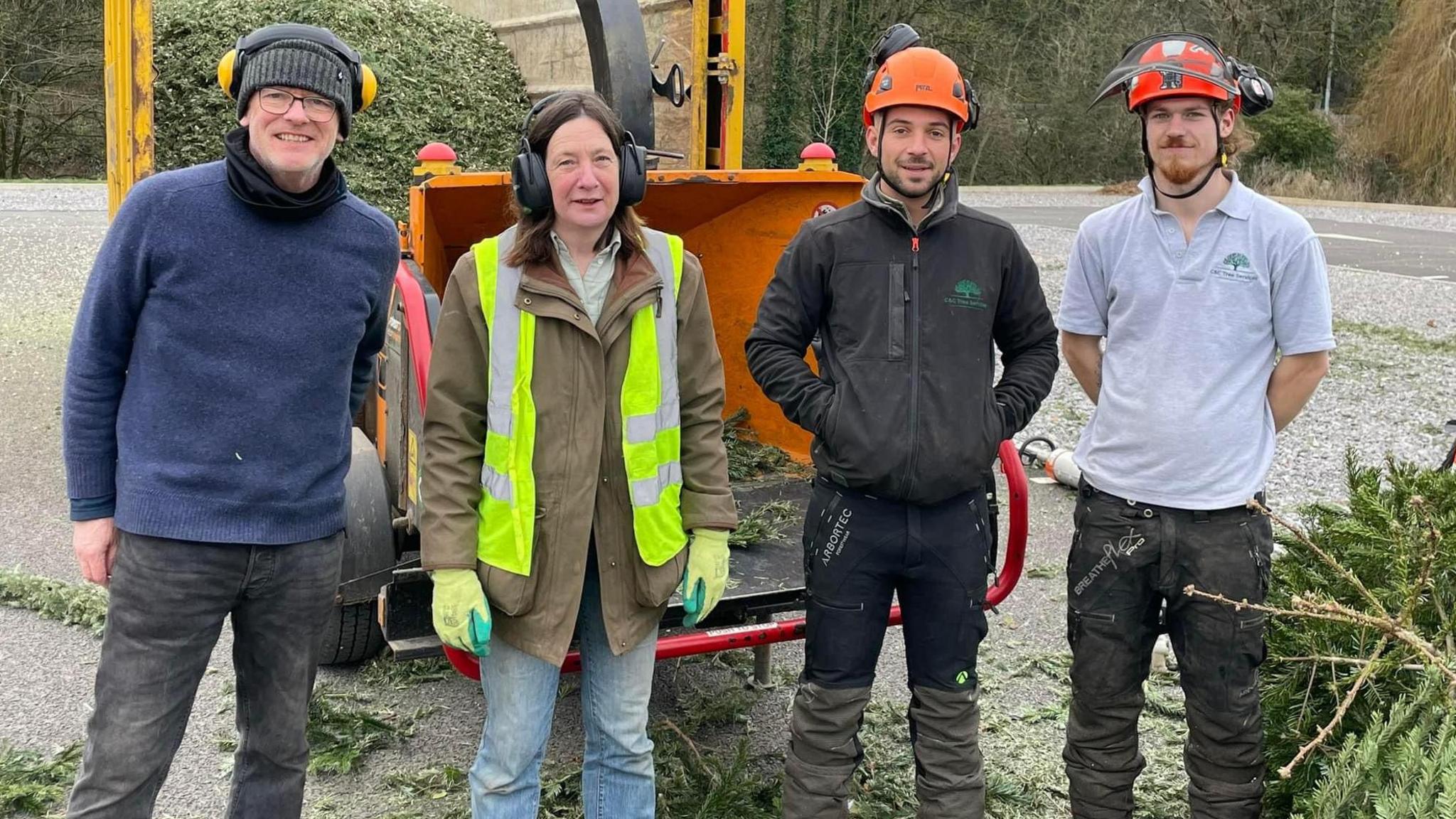 Four volunteers collecting old Christmas trees to raise money for Dorothy House Hospice. They are wearing orange hard hats and a small pile of Christmas trees is visible at their feed, while behind them is a machine that shreds the trees