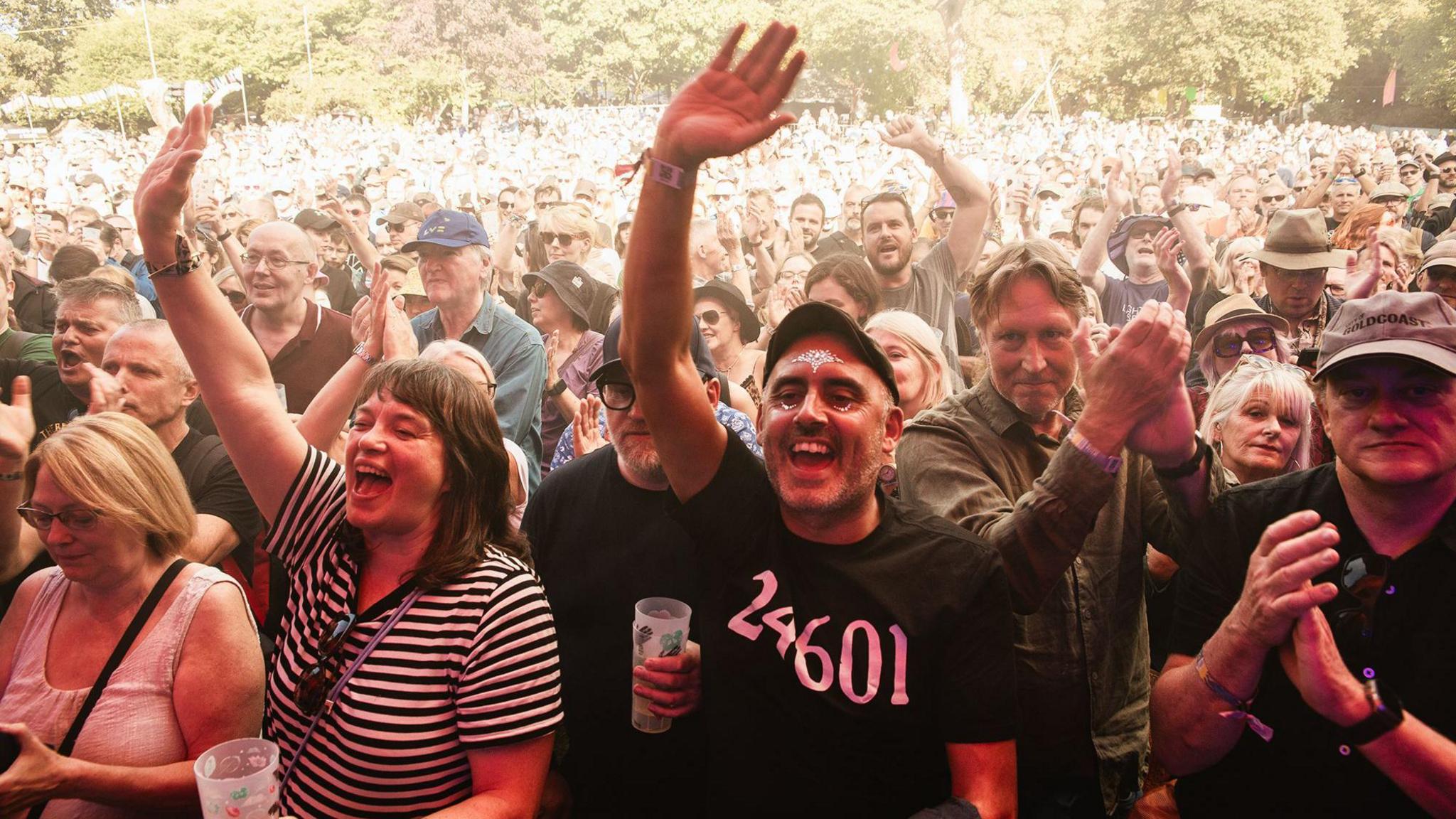 The crowd at Moseley Folk Festival, some of whom are holding pint glasses, and waving at the stage. A man in the forefront of the shot has a glitter tattoo on his forehead and cheeks. He and some others have arms aloft, others are clapping.