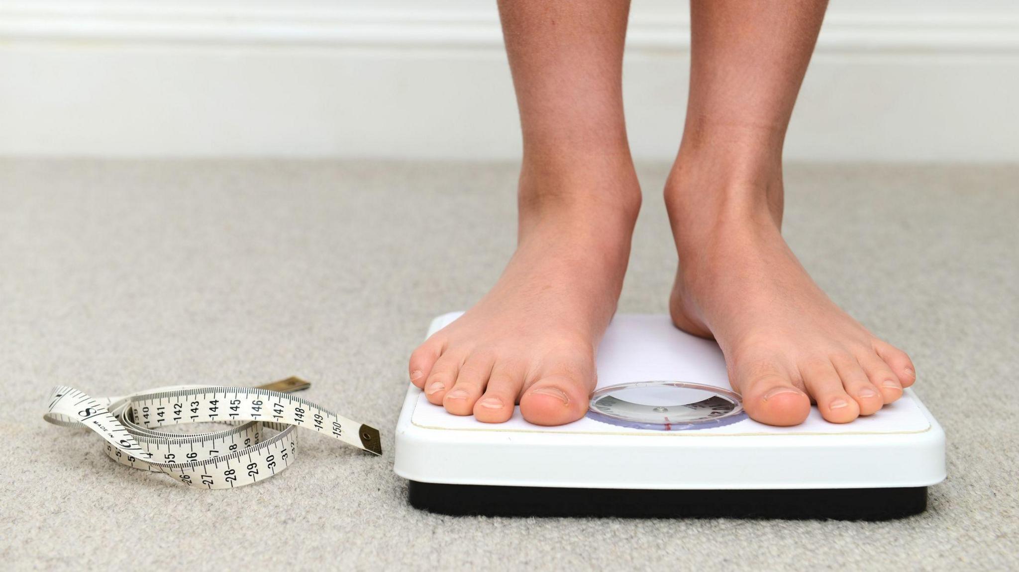 A child's feet standing on a set of scales, which are on a cream carpeted floor. The scales are white with a black base and have a dial in the centre. There is also a dressmaker's tapermeasure rolled up to the left of the scales