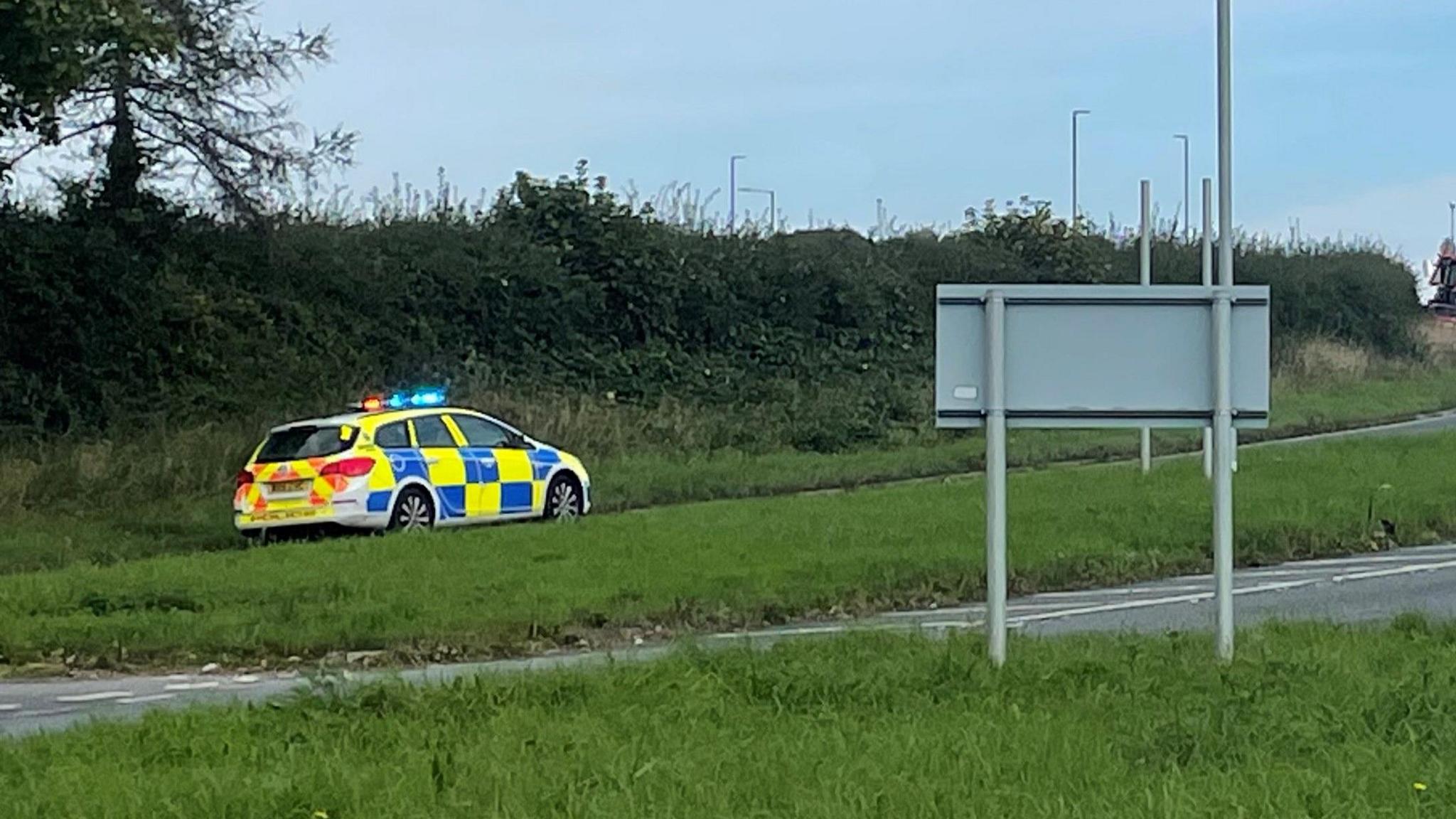 A police car with lights on, on the side of the road. A road sign on the right-hand side. 