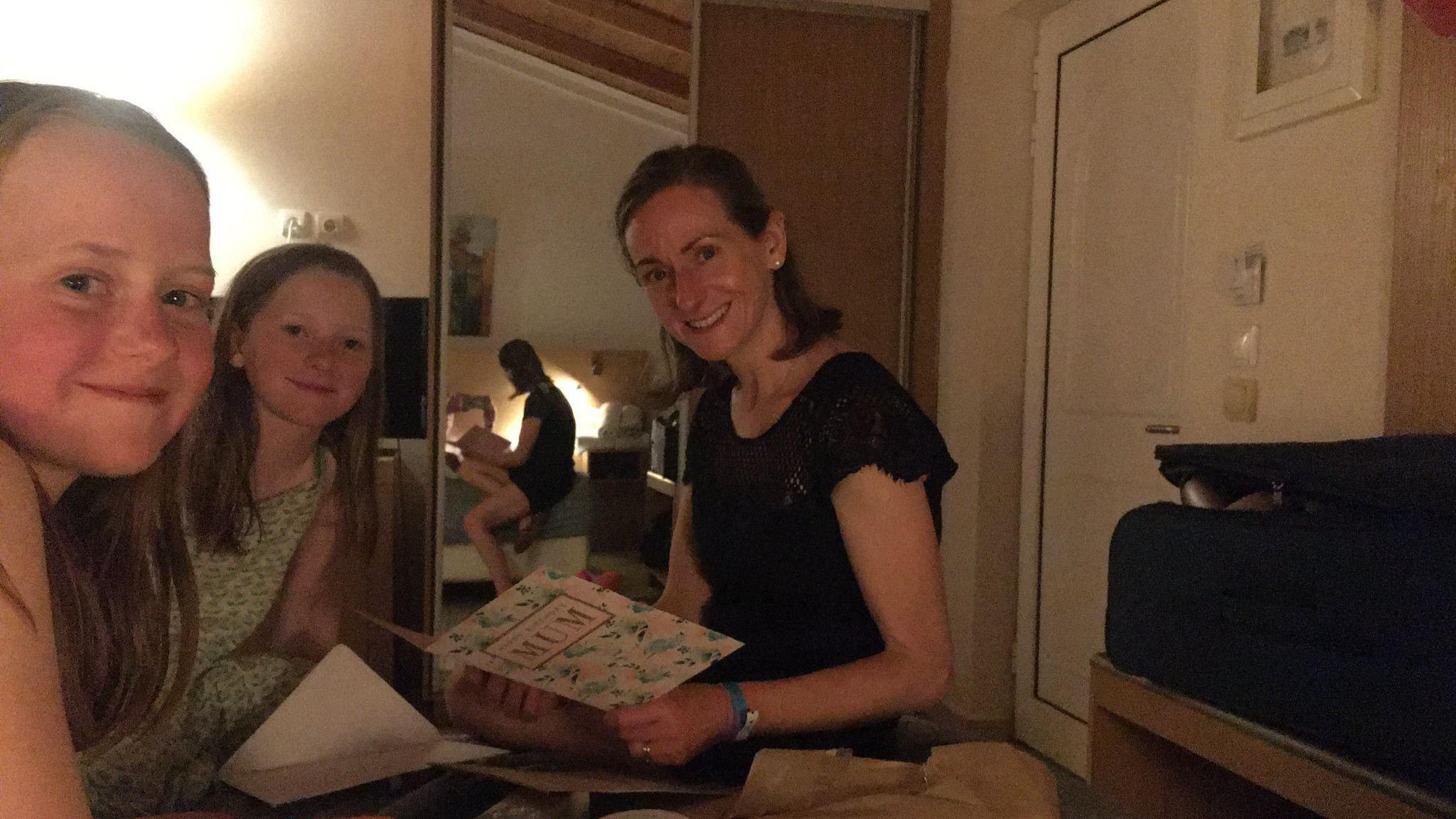 A woman is sat on a bed, holding a card that reads "mum" on the front. She is sat with two girls. All three are smiling whilst posing for the photograph. 