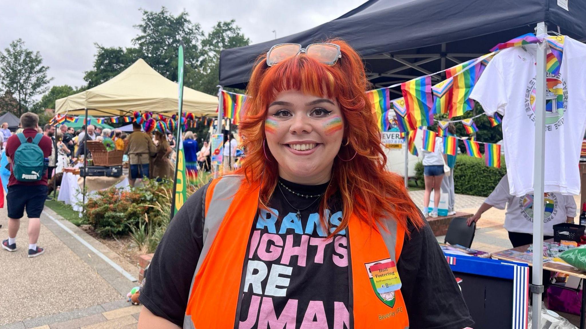 A head and shoulders photo of Elise Gilbert. She is wearing a black top with blue and pink bold writing on and an orange hi-visibility vest on. She has short red hair with a fringe and has blue, yellow and orange stripes pained on both of her cheeks. She is stood in front of a busy area with multiple canopy's and Pride flags. People can be seen in the background. The sky is overcast.