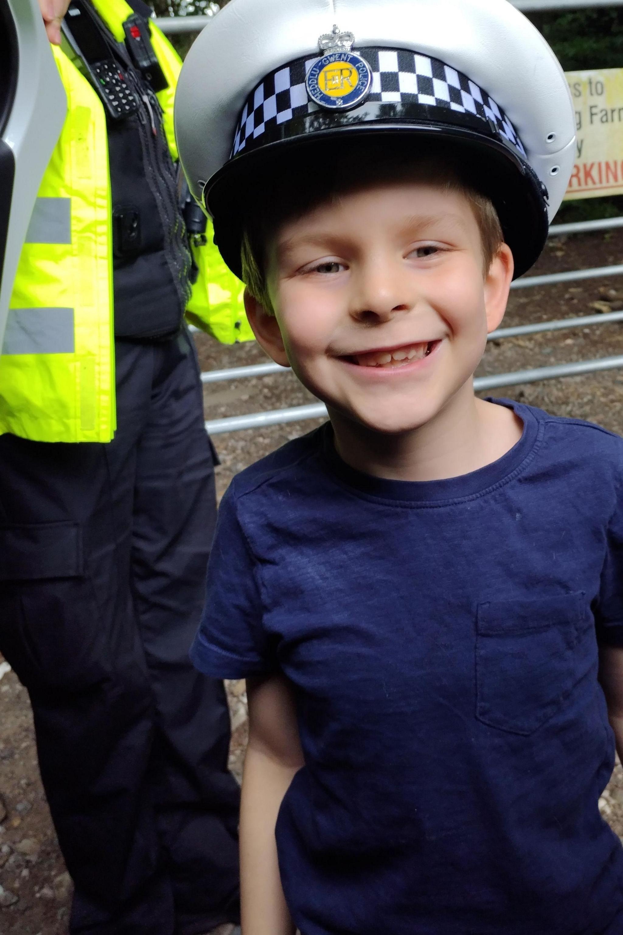 Macsen wearing a police hat and smiling at the camera with an officer stood behind him