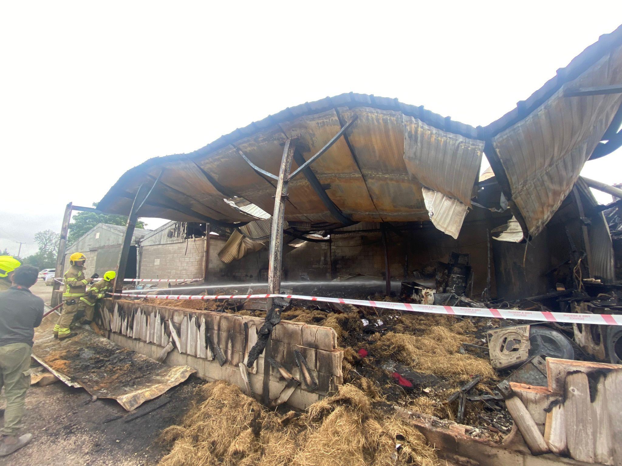 Firefighters are shown inspecting the remains of a large barn which has been badly damaged by fire. The corrugated iron roof has buckled and the area is cordoned off by red and white warning tape
