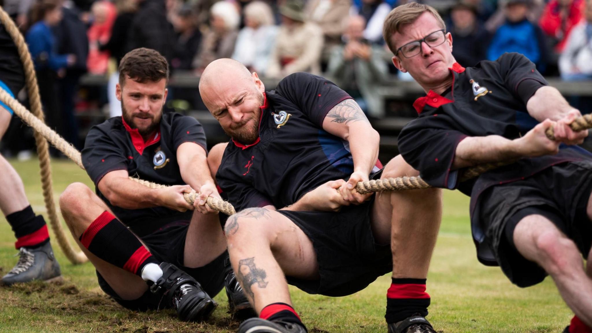 Three men taking part in a tug-of-war competition. They are pulling on a thick rope with grimacing faces. They are dressed in matching black uniforms with red collars and red socks.