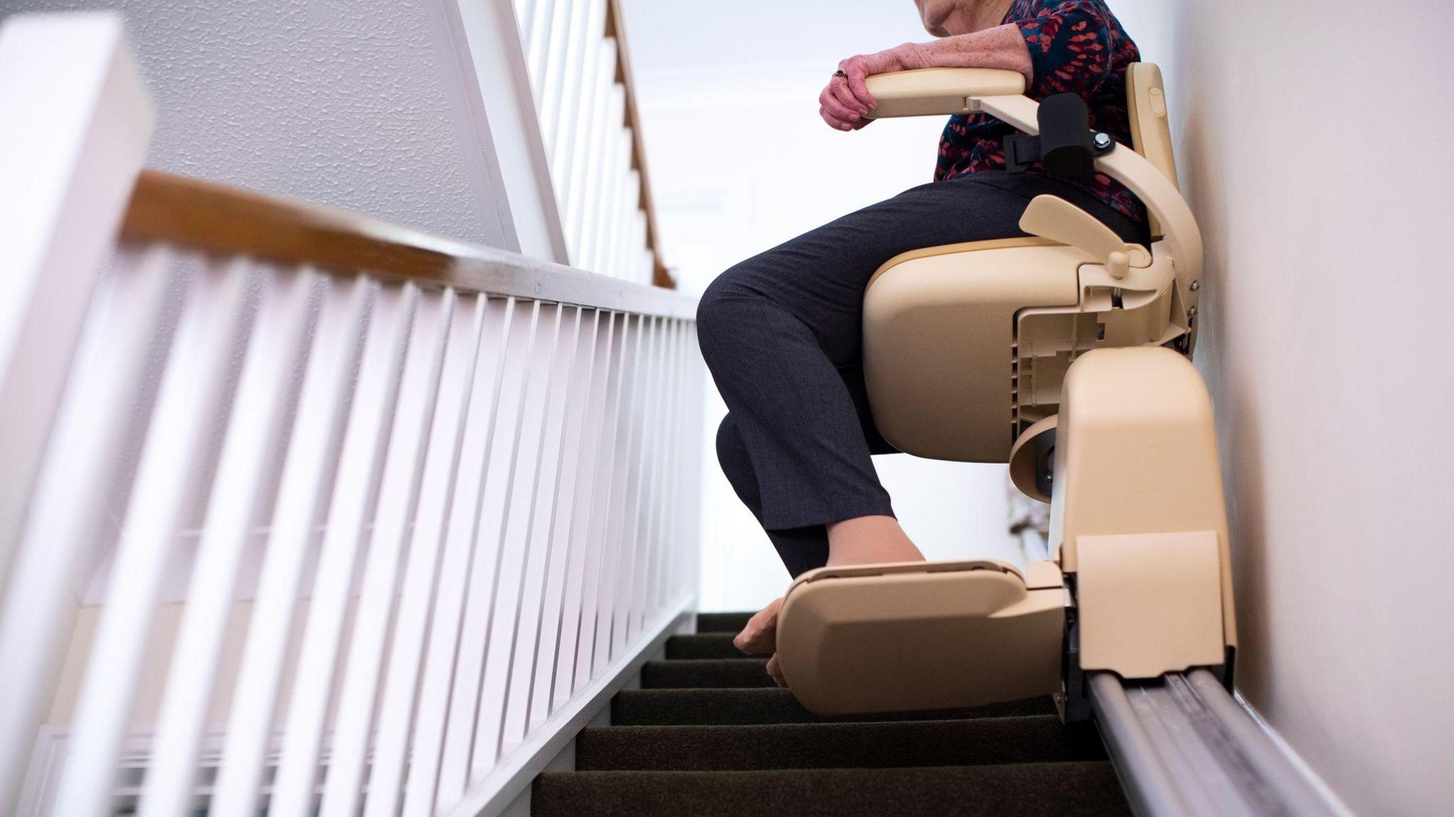 A woman sits in a stairlift which is going upwards. Only her legs and feet are visible. She is wearing blue trousers and sheer socks.
