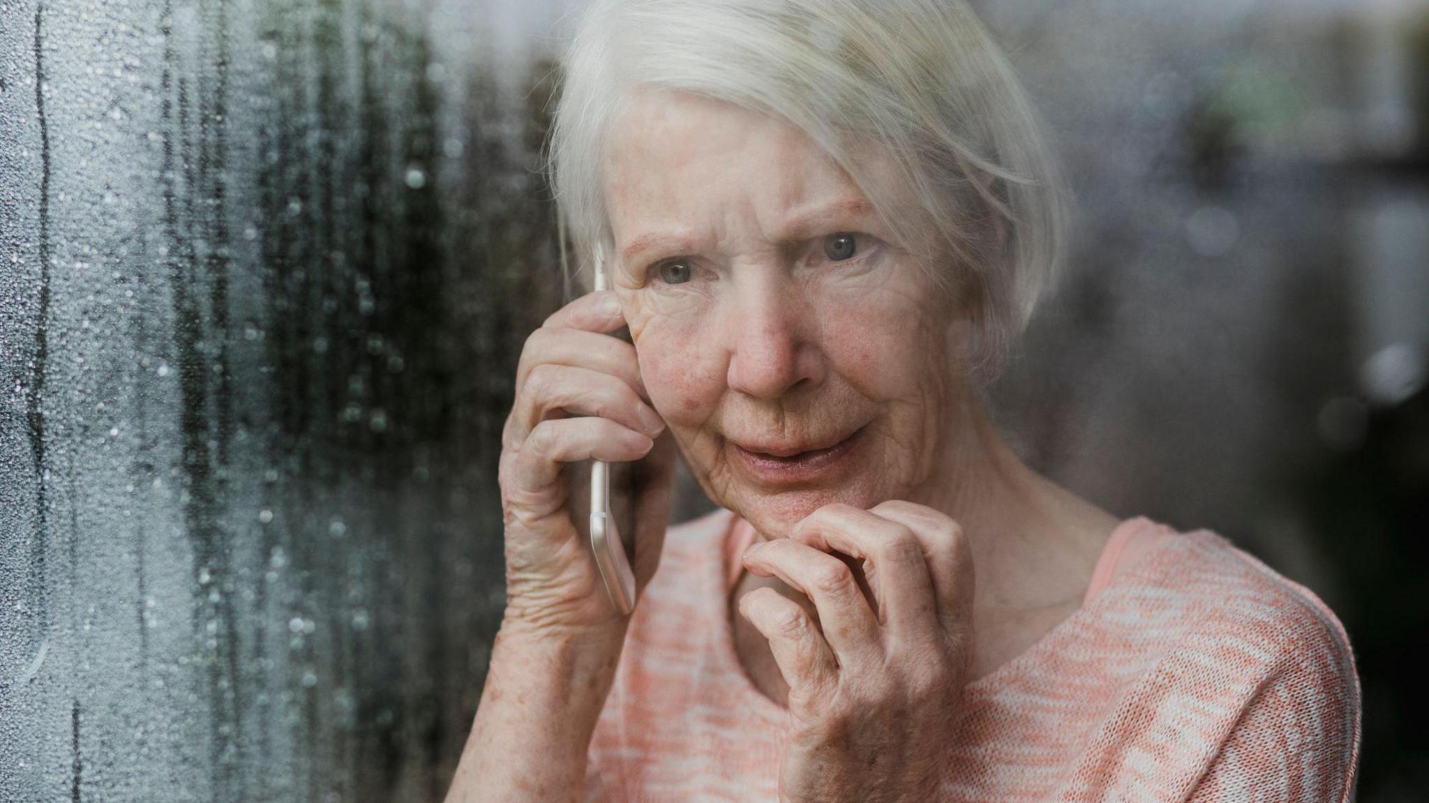 An older woman with white hair cut into a bob, wearing a salmon coloured top. She is on the phone while looking out through a window with rain trickling down it. Her facial expression looks slightly concerned.