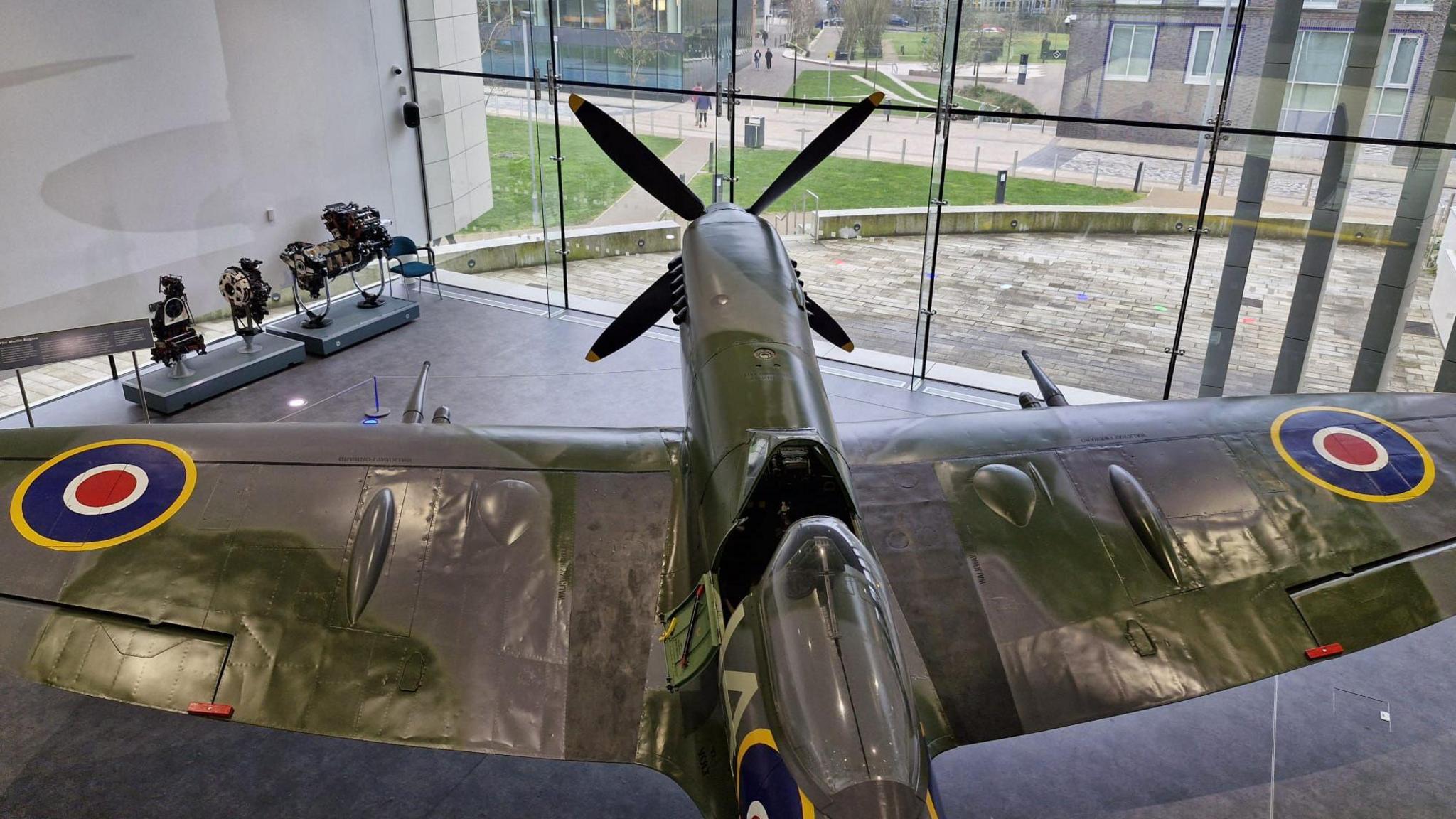 A Spitfire plane, viewed from above, is in a large glass-fronted building that also contains mechanical parts of the aircraft on display.