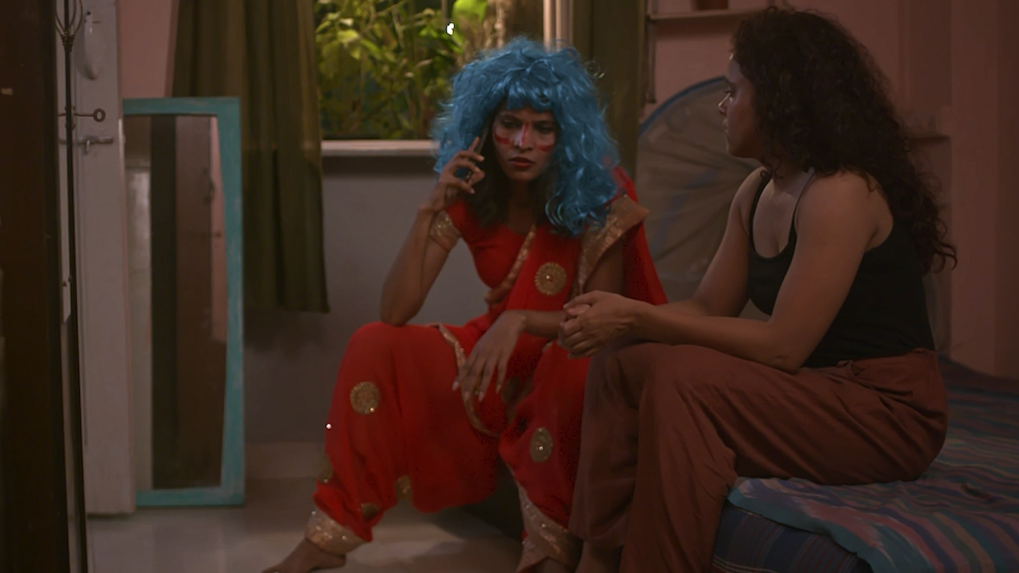 Two women in a dimly lit apartment - one wearing a black vest, the other wearing an ornamental red saree with a blue wig, talking on the phone.