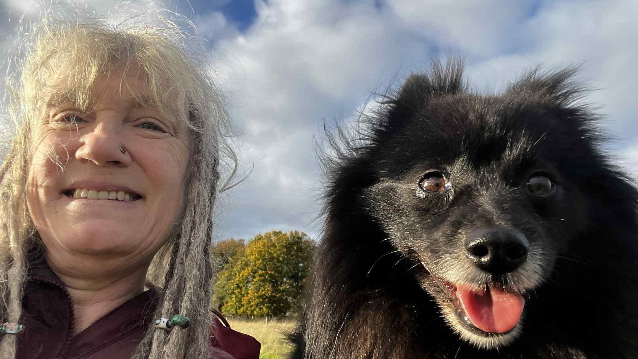 A close up selfie of Dee with her dog Bilbo. Dee has white blonde hair with dreadlocks. Next to her is Bilbo, a small black dog which smiles at the camera. He has fine, straight black hair and dark brown eyes. They're sat in a field whilst on a dog walk.