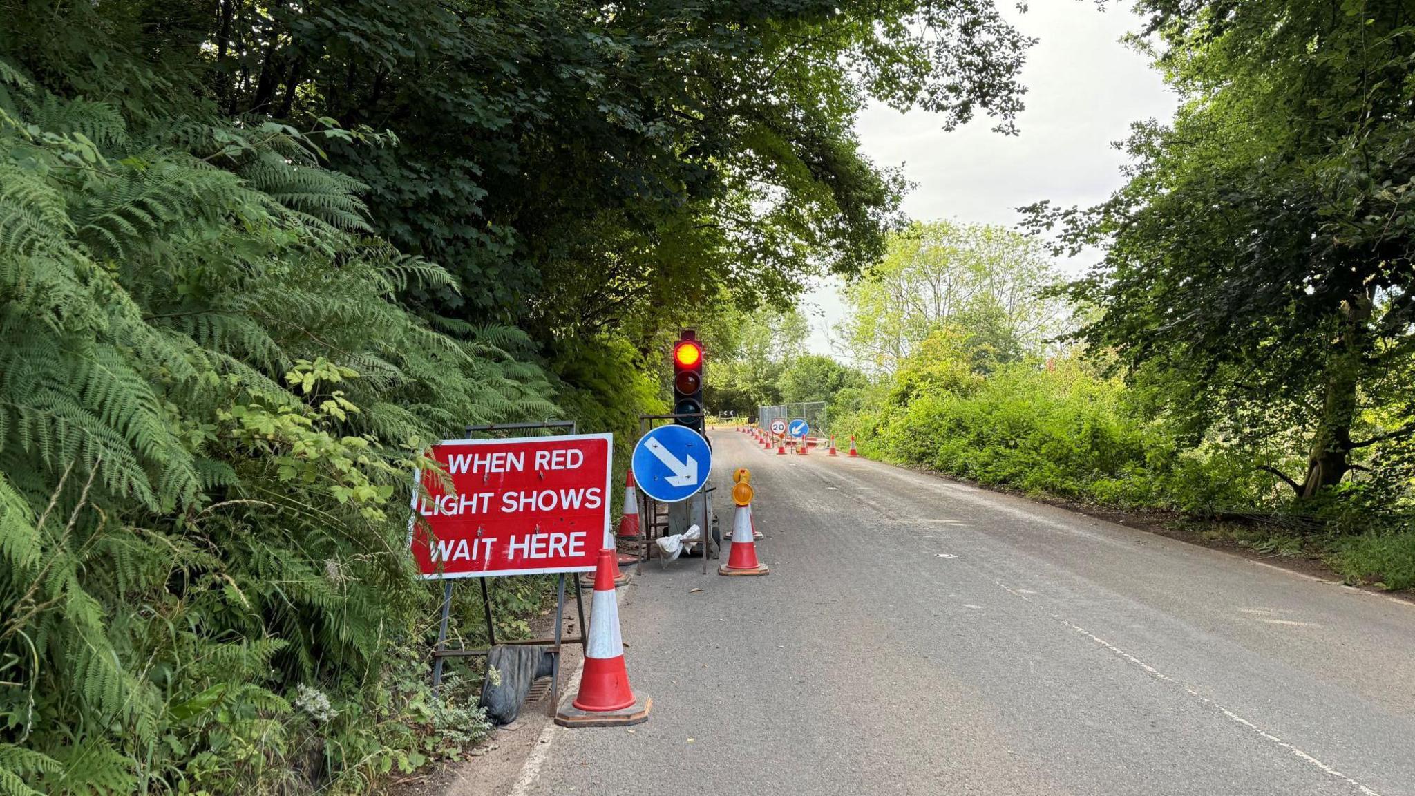A sign with white letters on a red background reads "when red light shows wait here" on a rural road near Wheddon Cross in Somerset. There are trees and bushes lining each side of the road, which is single lane in each direction. There are also traffic cones and a set of traffic lights visible