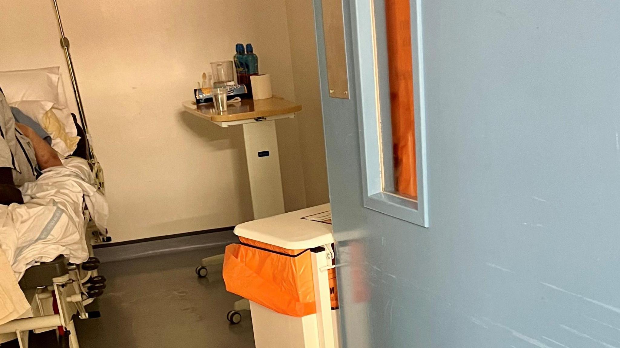 The right hand side of a man is to the left of the frame, he lies on a hospital bed. Beside the blue door is a white medical bin with a orange bin liner. On the table beside him  is some juice and biscuits