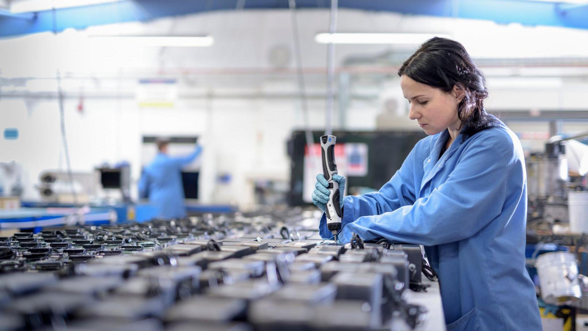 A young woman wearing a blue boilersuit uses a tool on components sitting on a benchtop.