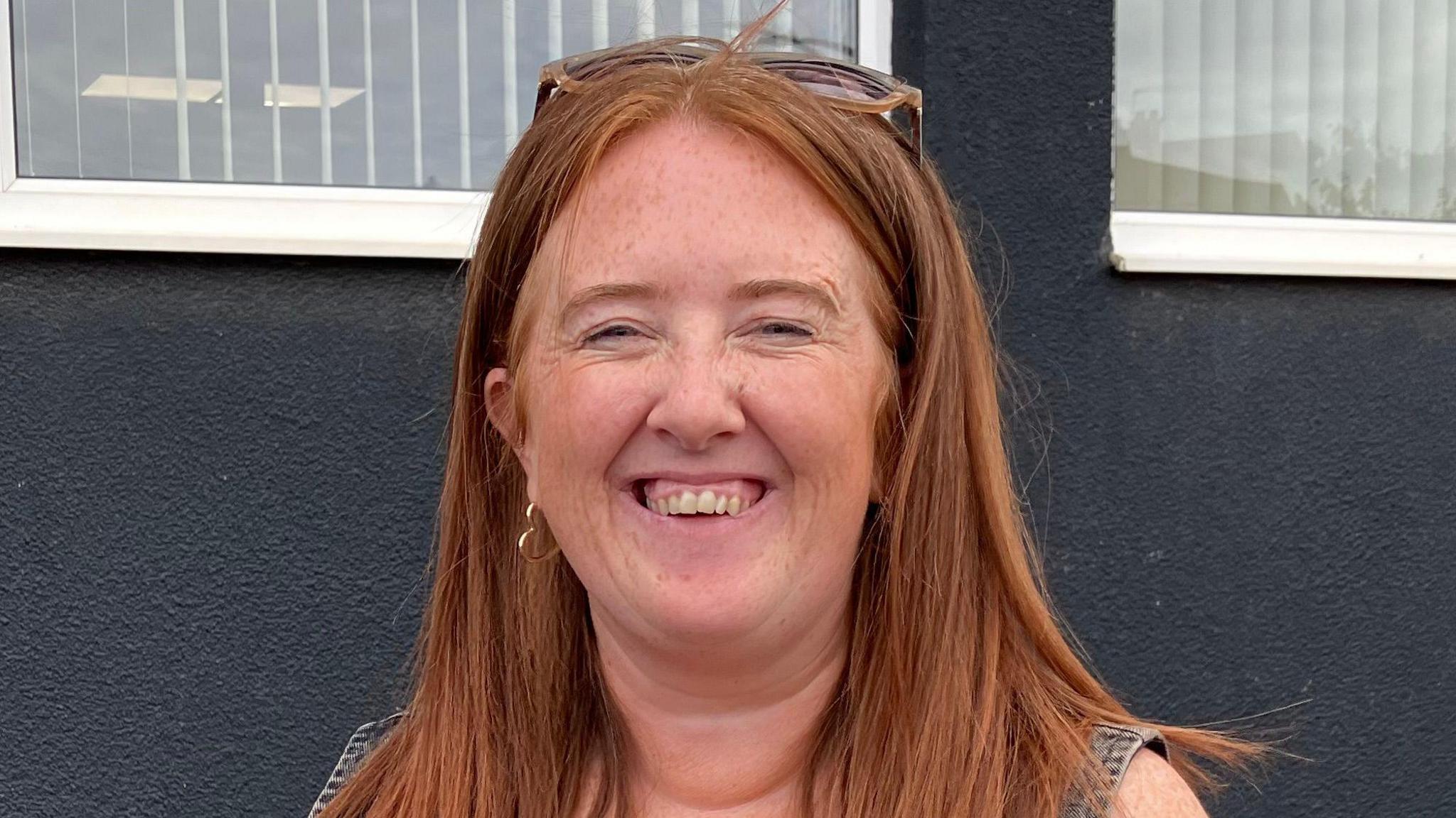 A woman with long ginger hair smiles at the camera. She is standing against a blue wall.