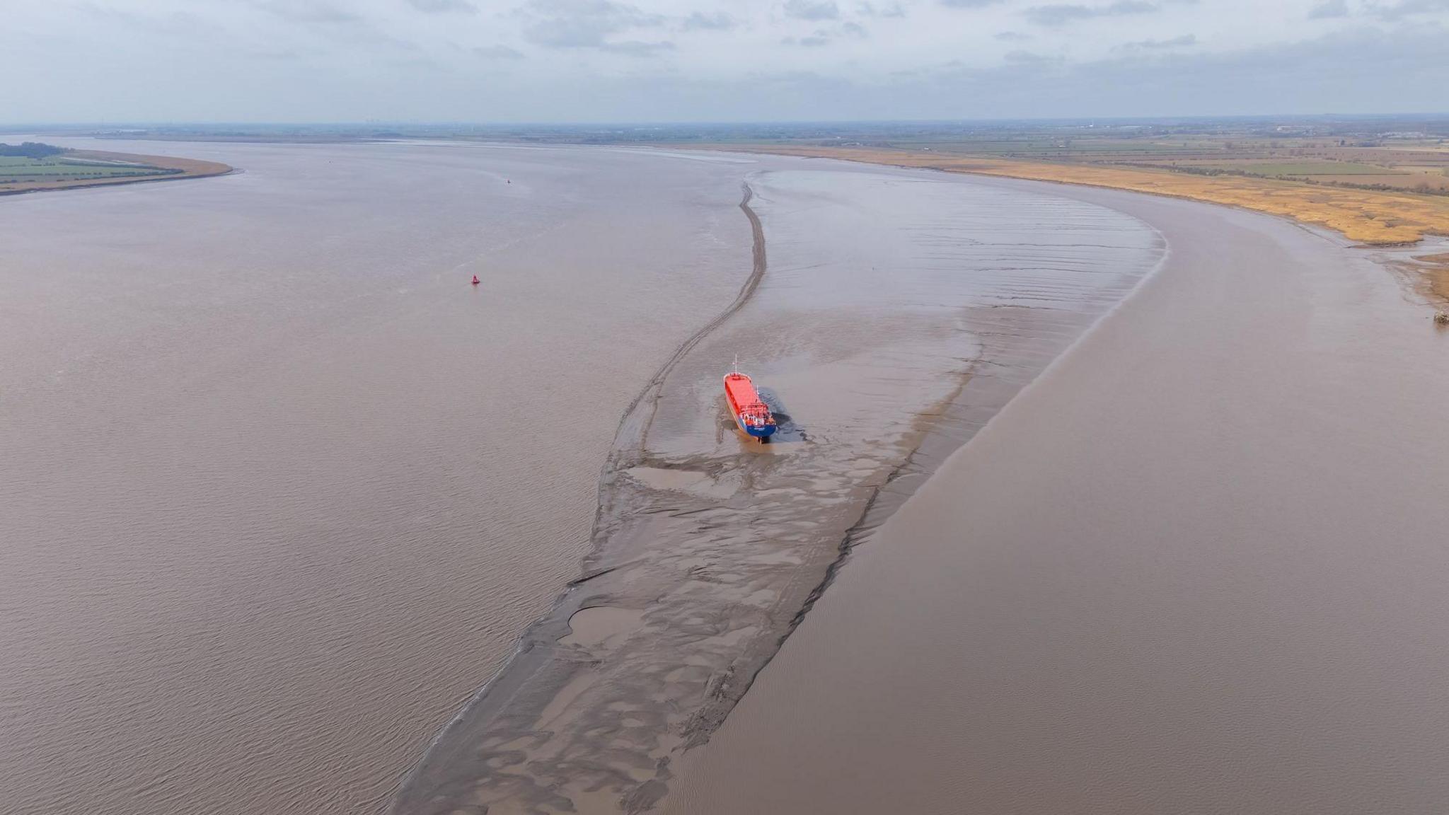 A red and blue cargo ship stuck in mud on the Humber. The photo is taken from a high aerial perspective and the ship is in the distance with a vast amount of water surrounding it.