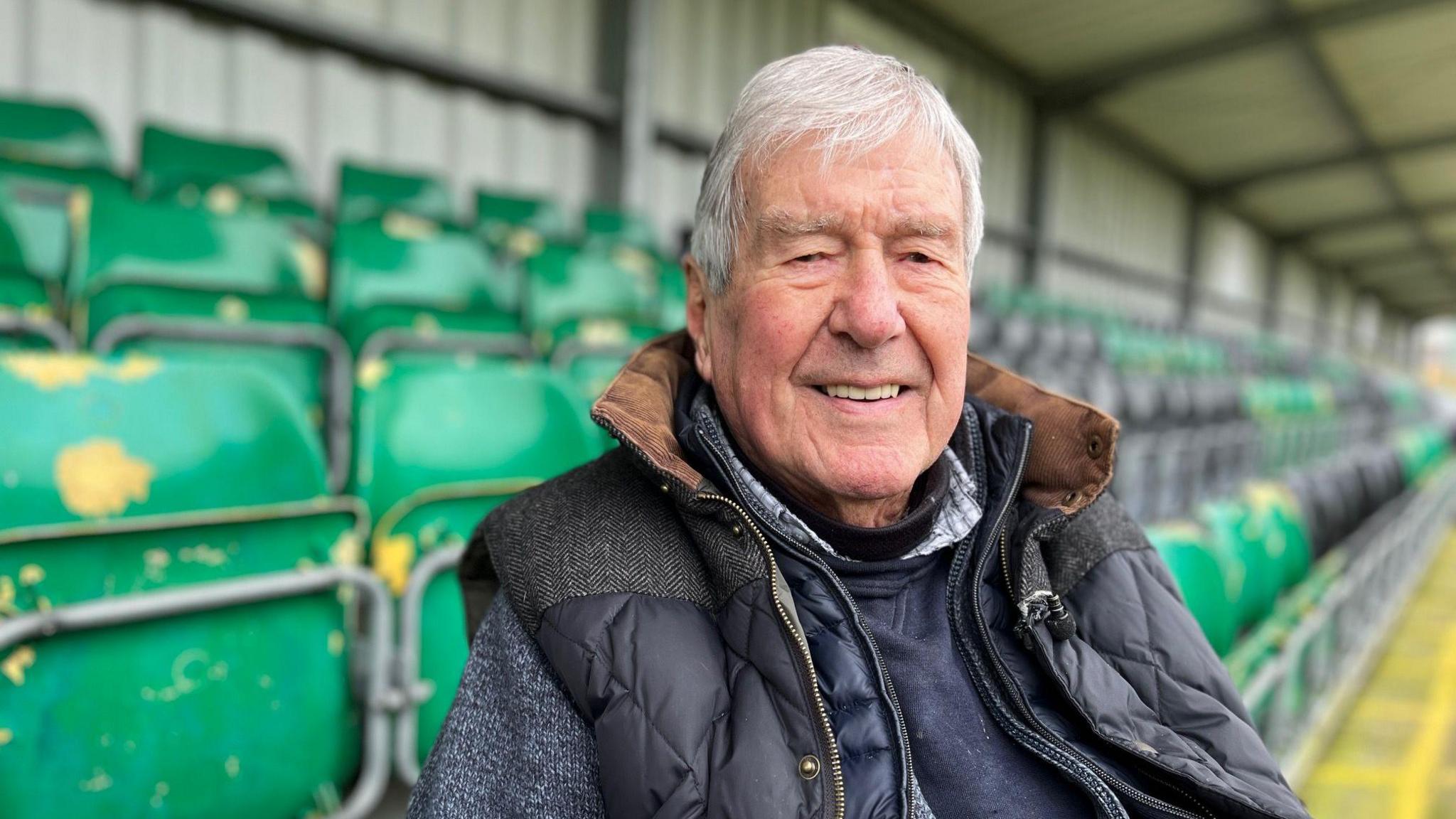 An elderly man with white hair sits on a green chair in a football stadium and smiles at the camera. 