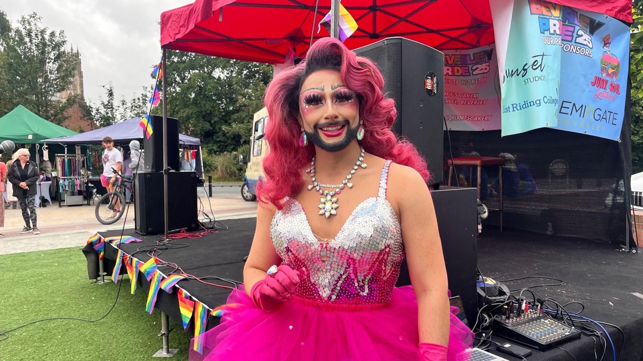 Drag artist Queen Margarita stood in front of a stage which has a red canopy and is lined with Pride flags. Queen Margarita has long pink hair, a black moustache and beard and is wearing colourful earrings, a silver nicklace, pink gloves, a silver strappy top and a pink tutu. 