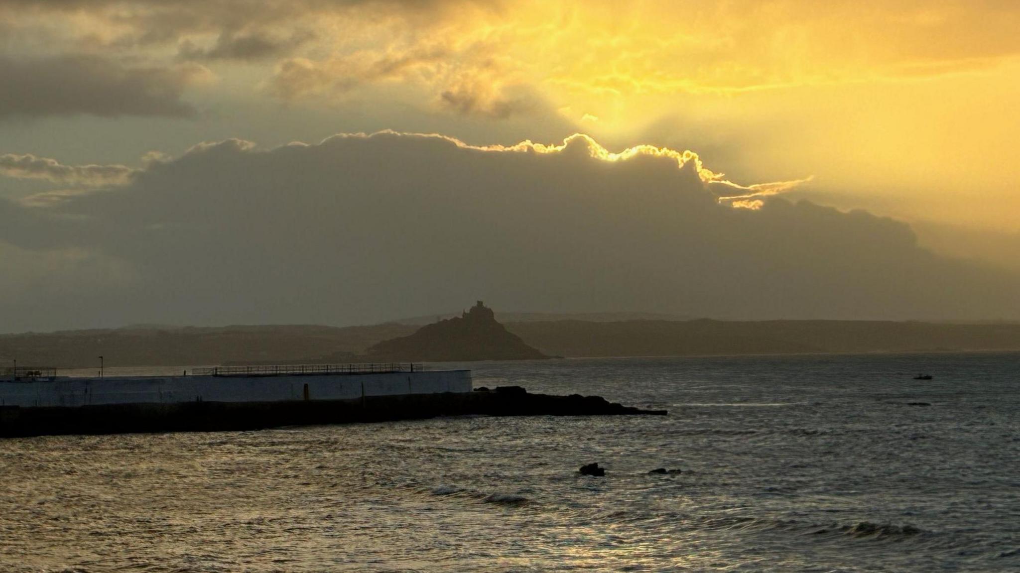 A picture of a large body of water, with an island in the distance. Above the water, the yellow glow of the sun can be seen breaking through the clouds.