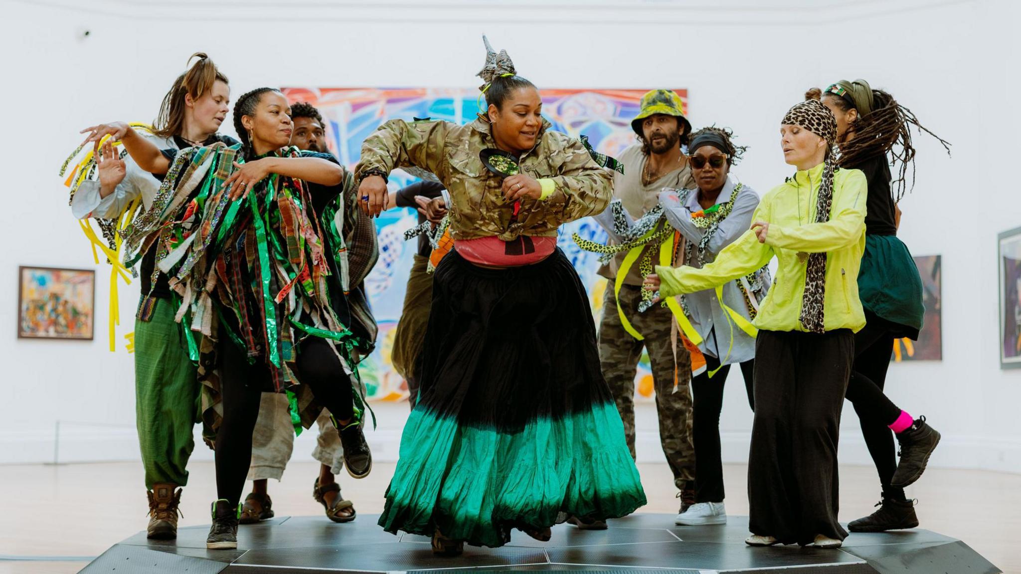 A group of eight dancers mid-movement. A woman centre of the group is wearing a khaki long sleeved top with a black and green long skirt. 
Other dancers are wearing a mix of outfits, with one woman in a lime green top and black trousers and others with green and brown long ribbon-style material over their shoulders. They are wearing boots and trainers and all look like they are enjoying the dance.