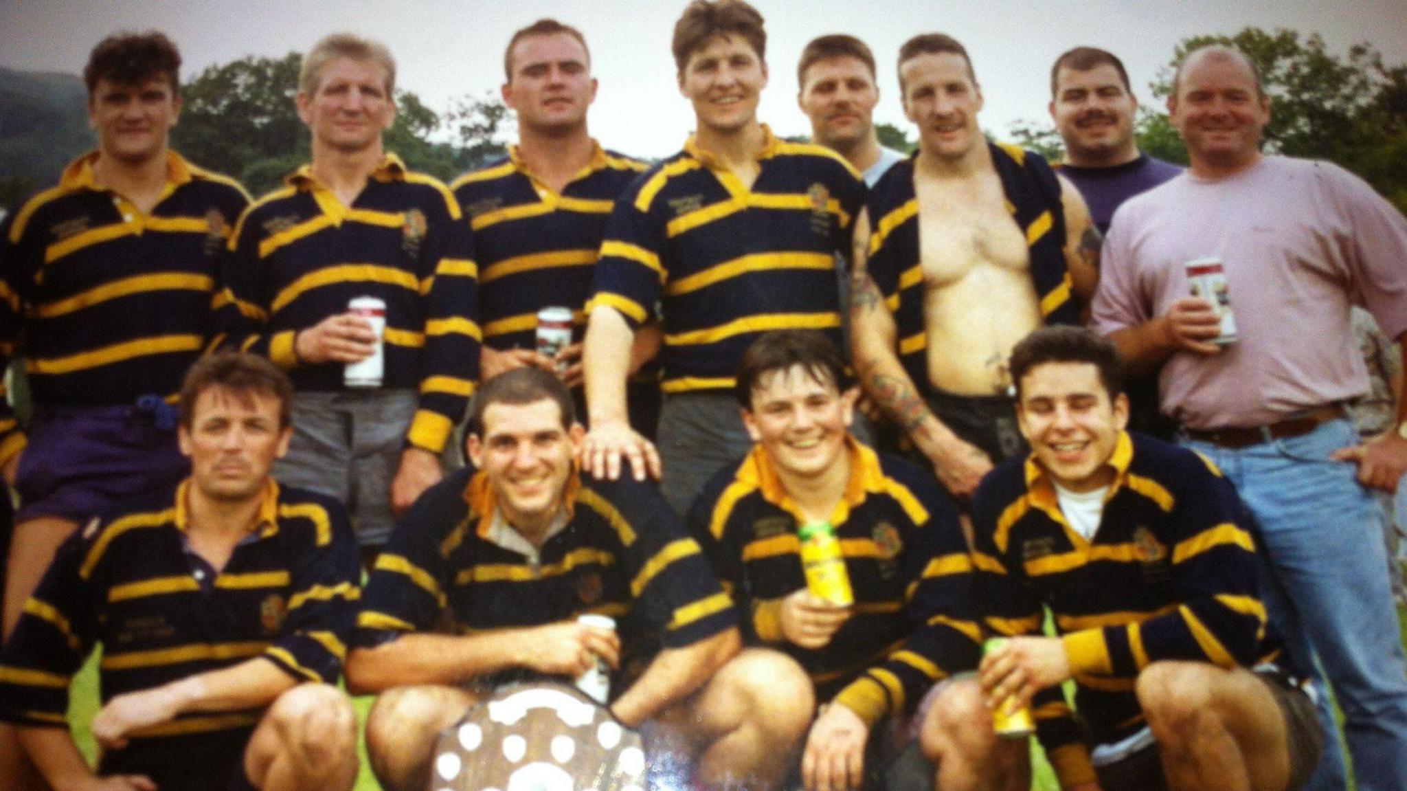 Nick Servini with his rugby team mates when he was younger. He is wearing Aberdare RFC black and yellow striped kit and holding a can of beer. The team is standing in two rows with one player in the middle of the bottom row holding a prize shield. 