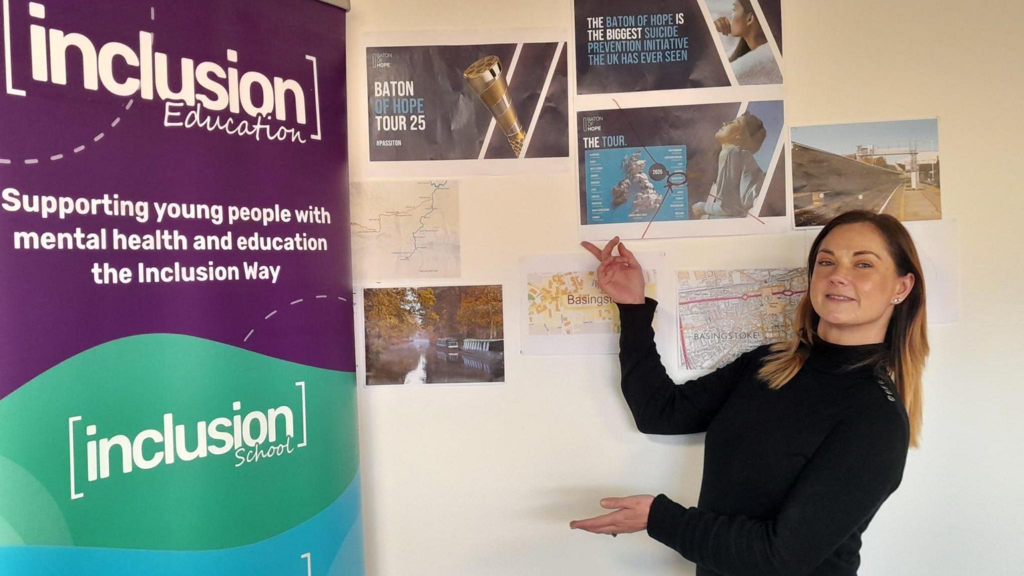 Marie Greenhalgh pointing to a sign that says: Inclusion Education: supporting you people with mental health and education the Inclusion Way. There are also pieces of paper promotion the Baton of Hope Tour 2025 behind her on a wall