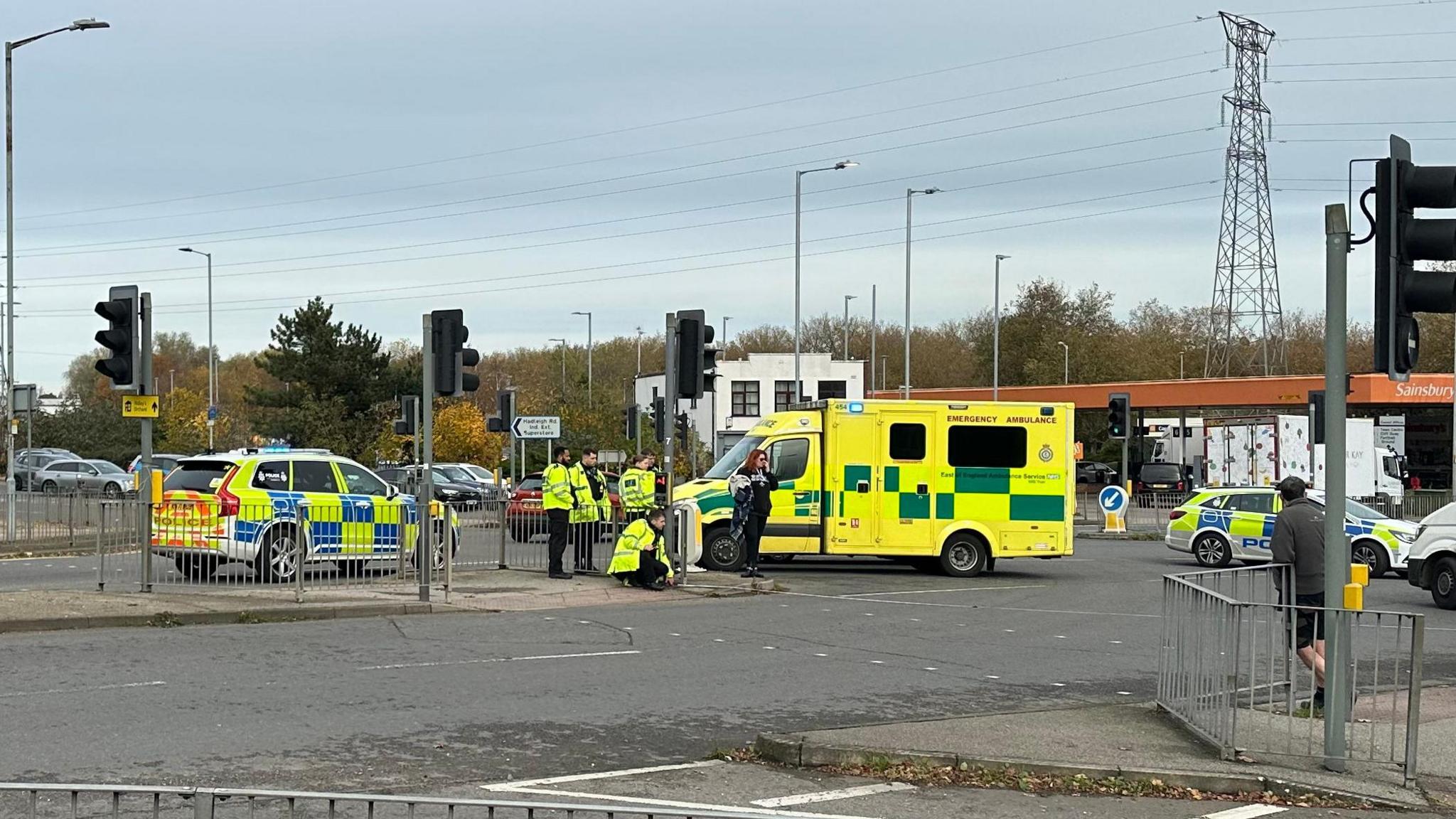 Another view of the emergency services at the scene of the collision. Officers can be seen in hi-vis jackets investigating the road. An ambulance is also parked nearby. Pedestrians can be seen moving about the area around the scene.