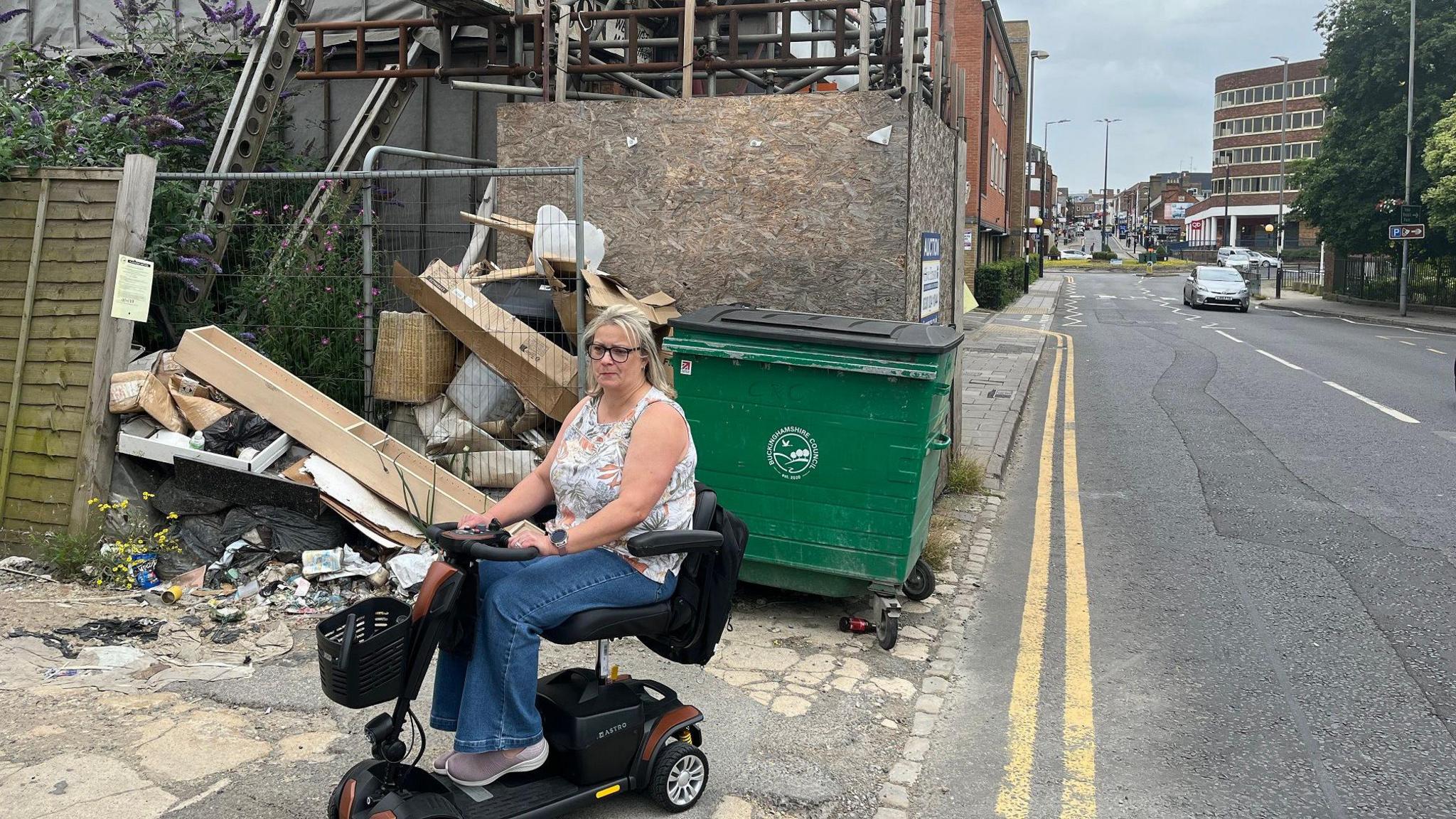 Lynda Bell sat on a mobility scooter in front of the scaffolding. She is right next to a road with a car travelling on it. She has blonde hair, glasses and is wearing a white top with blue jeans.