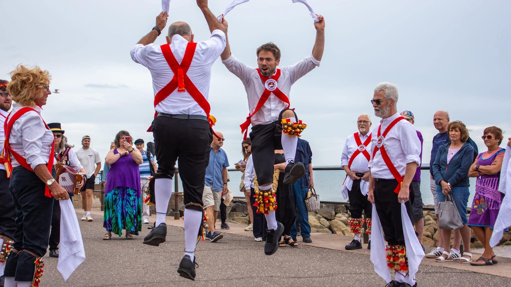 Two Morris dancers leap into the air while doing a performance along Sidmouth seafront during the town's Folk Festival,. They are wearing black trousers, white shirts, red sashes and red and yellow bells, which are on their legs. They have white handkerchiefs in their hands. Several people are stood watching the performance.