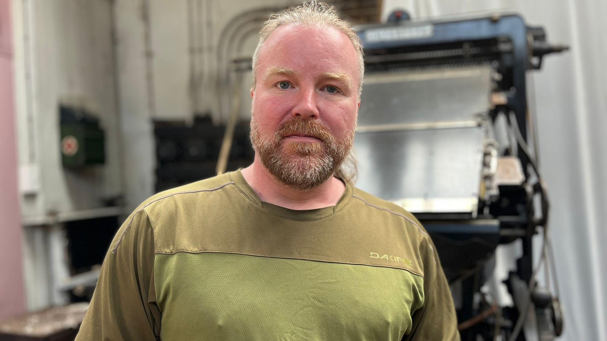 Graeme Berry stands in front of a printing press - he has light red hair and beard and wears an olive green t-shirt.