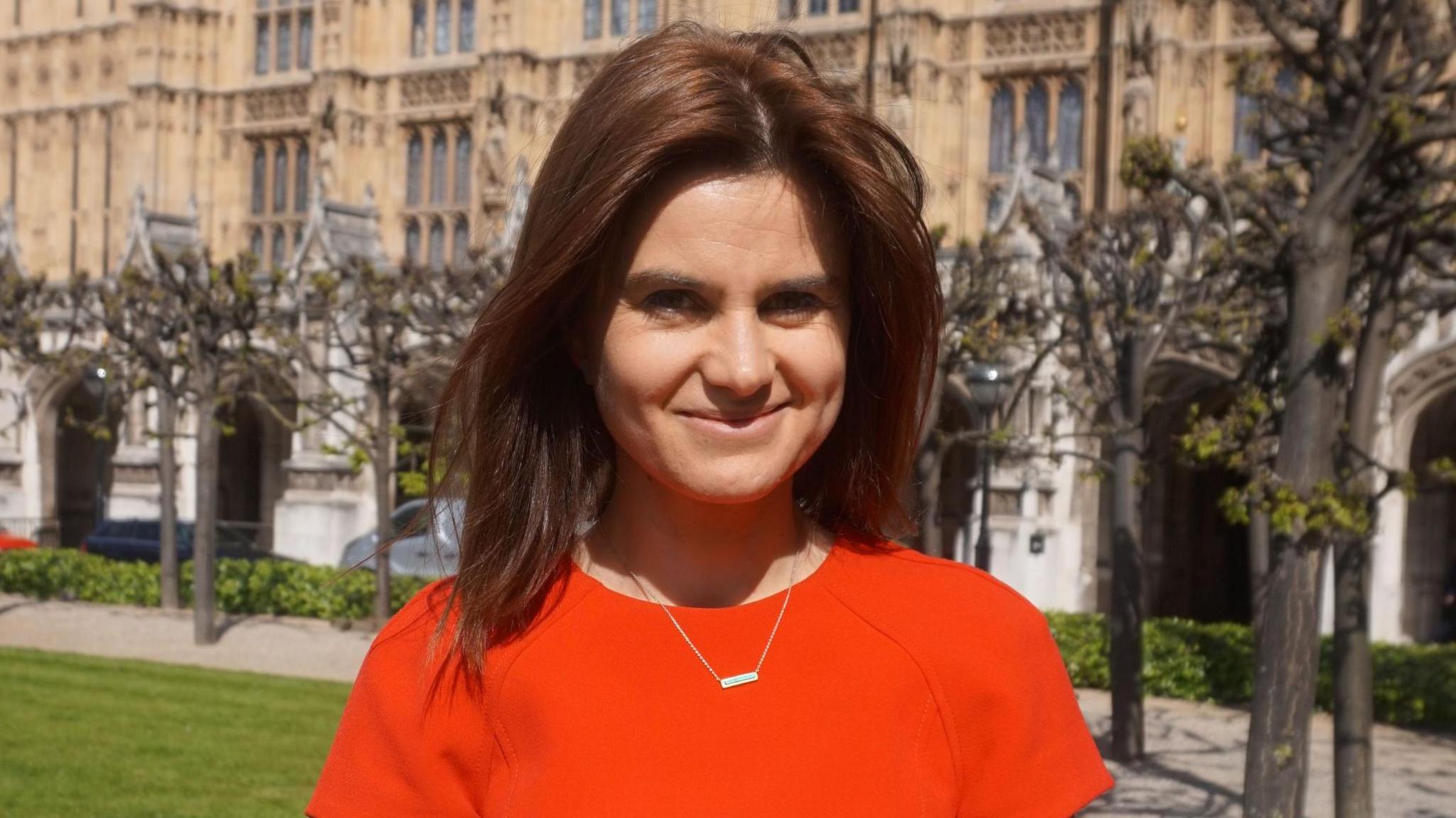 Jo Cox MP, a brunette woman, smiles at the camera. She wears an orange top with a necklace. Behind her the houses of parliament are blurred.