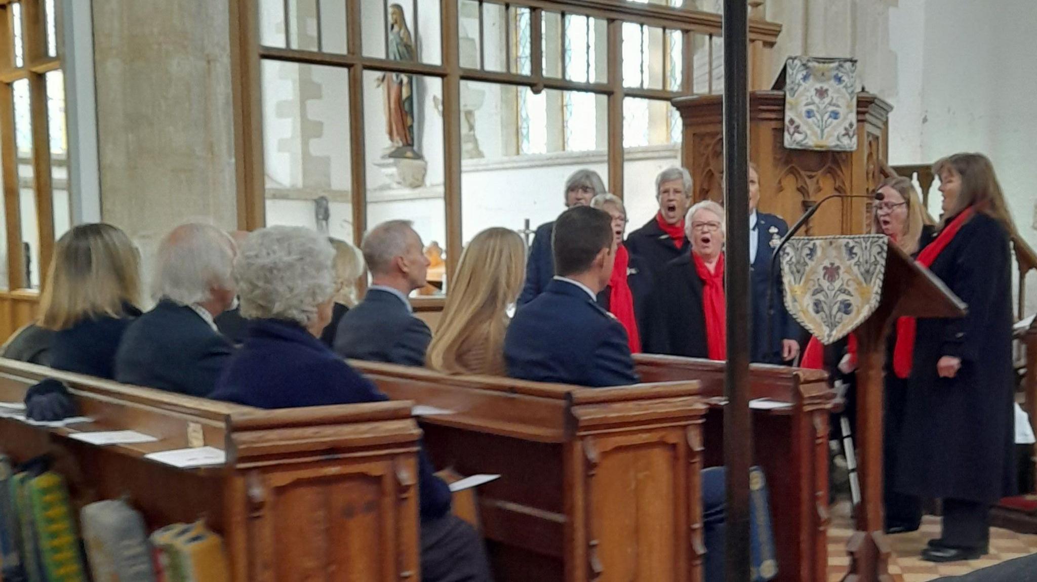 A choir sings. They wear black gowns with red scarves. members of the congregation sit in pews. 