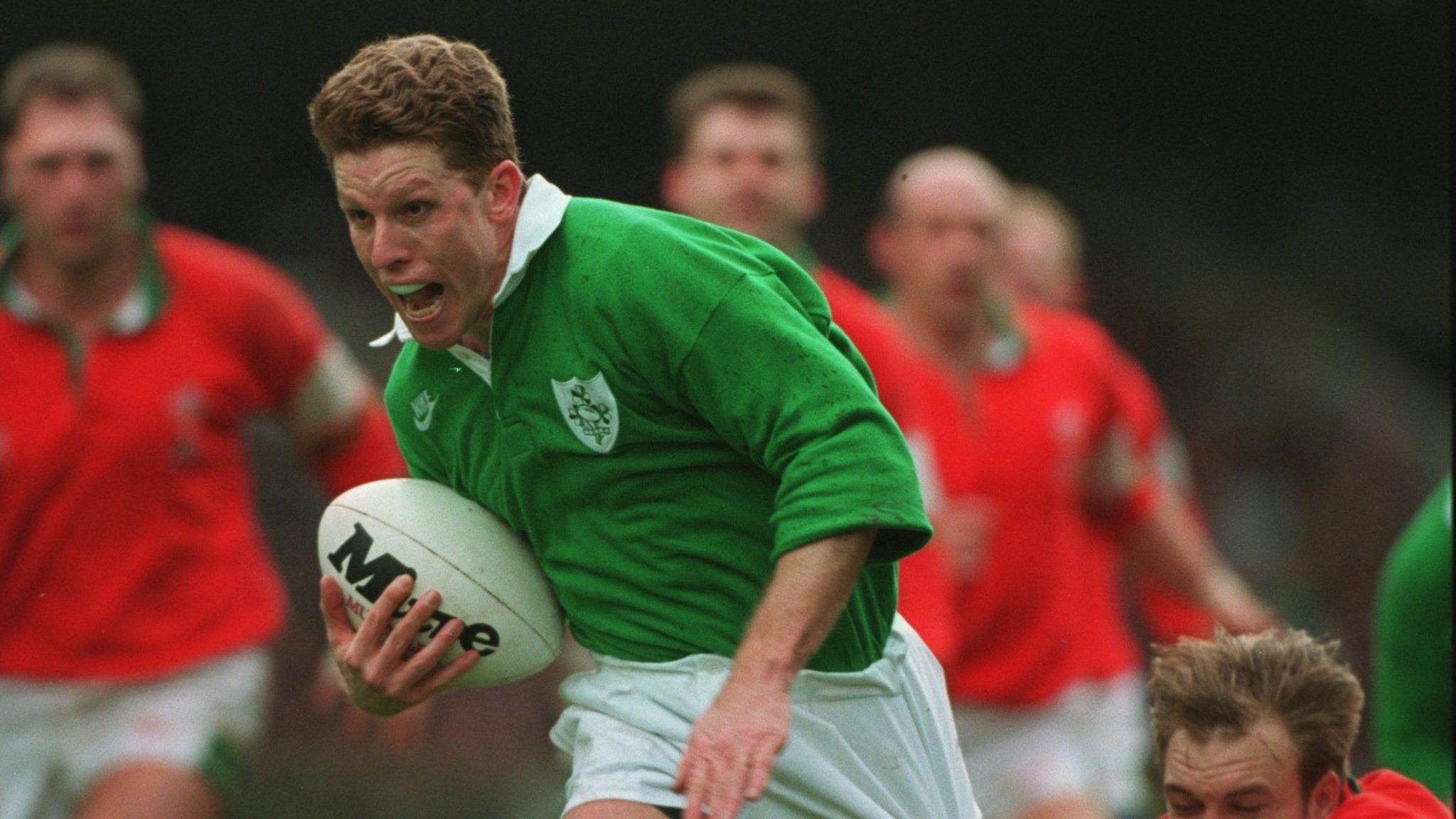 Mullin running with the ball as he plays rugby in 1995. He has thick brown hair and is wearing a green top and white shorts as he holds the ball in his right hand.