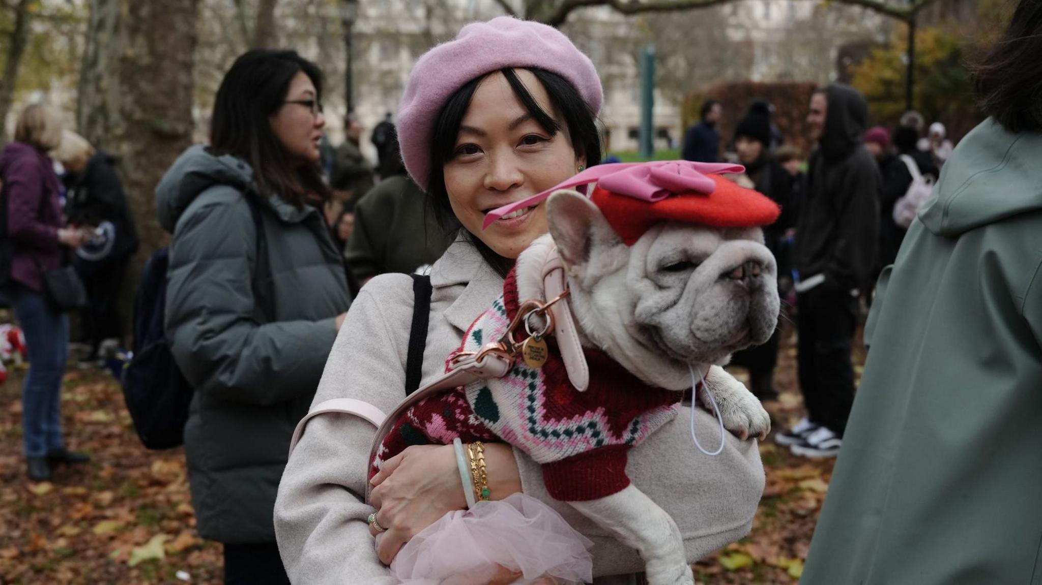 A woman in a grey coat and pink beret holds a dog with a red beret and a pink and red jumper.