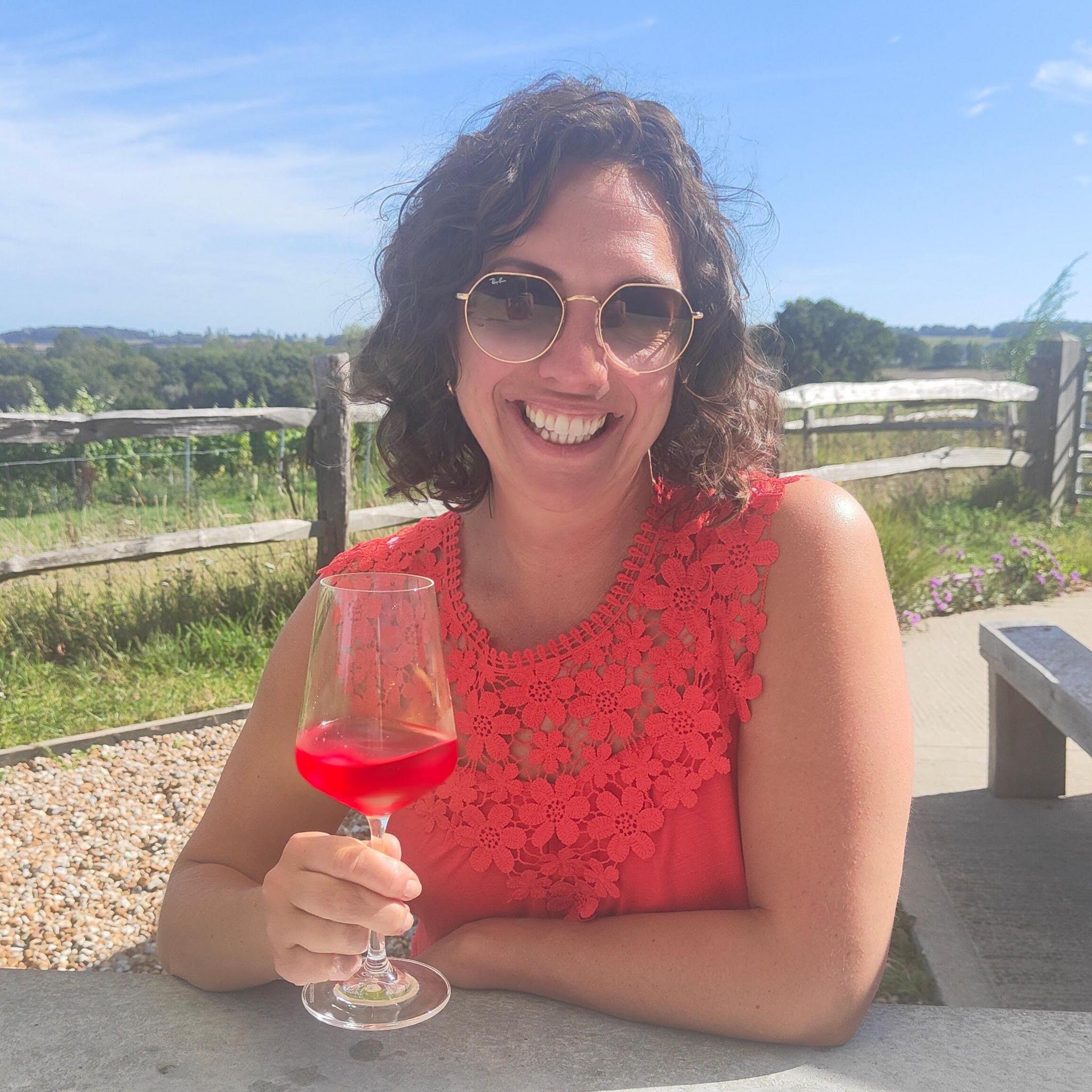 Wine glass containing a bright red drink held in the foreground by a person seated at an outdoor table. The person wears round sunglasses and a sleeveless top with a floral lace pattern. A wooden fence, gravel area, plants and open countryside are visible in the background.