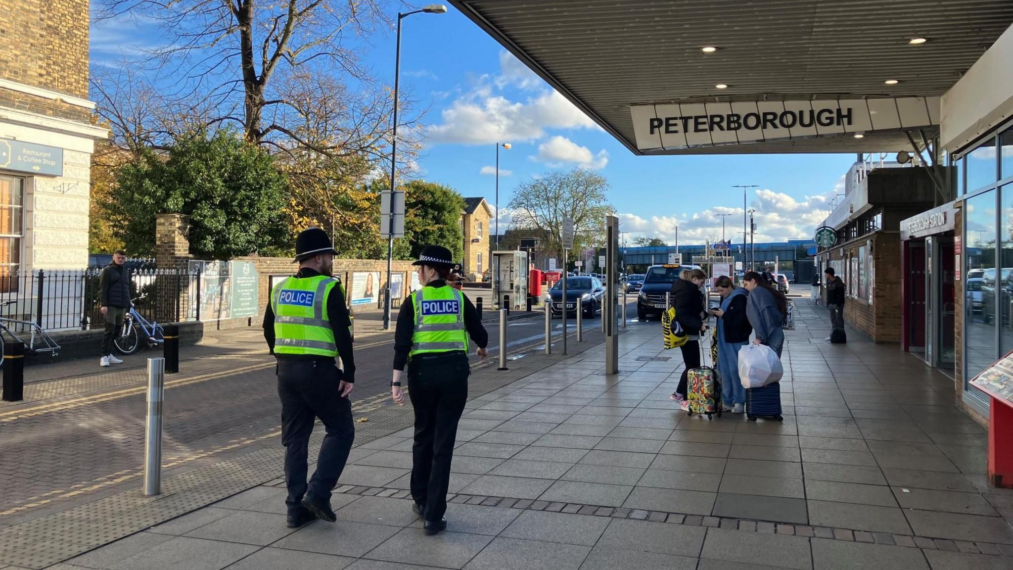 Two police officers in yellow tabards walking on the concourse outside Peterborough railway station. There are a handful of other people on the pavement.