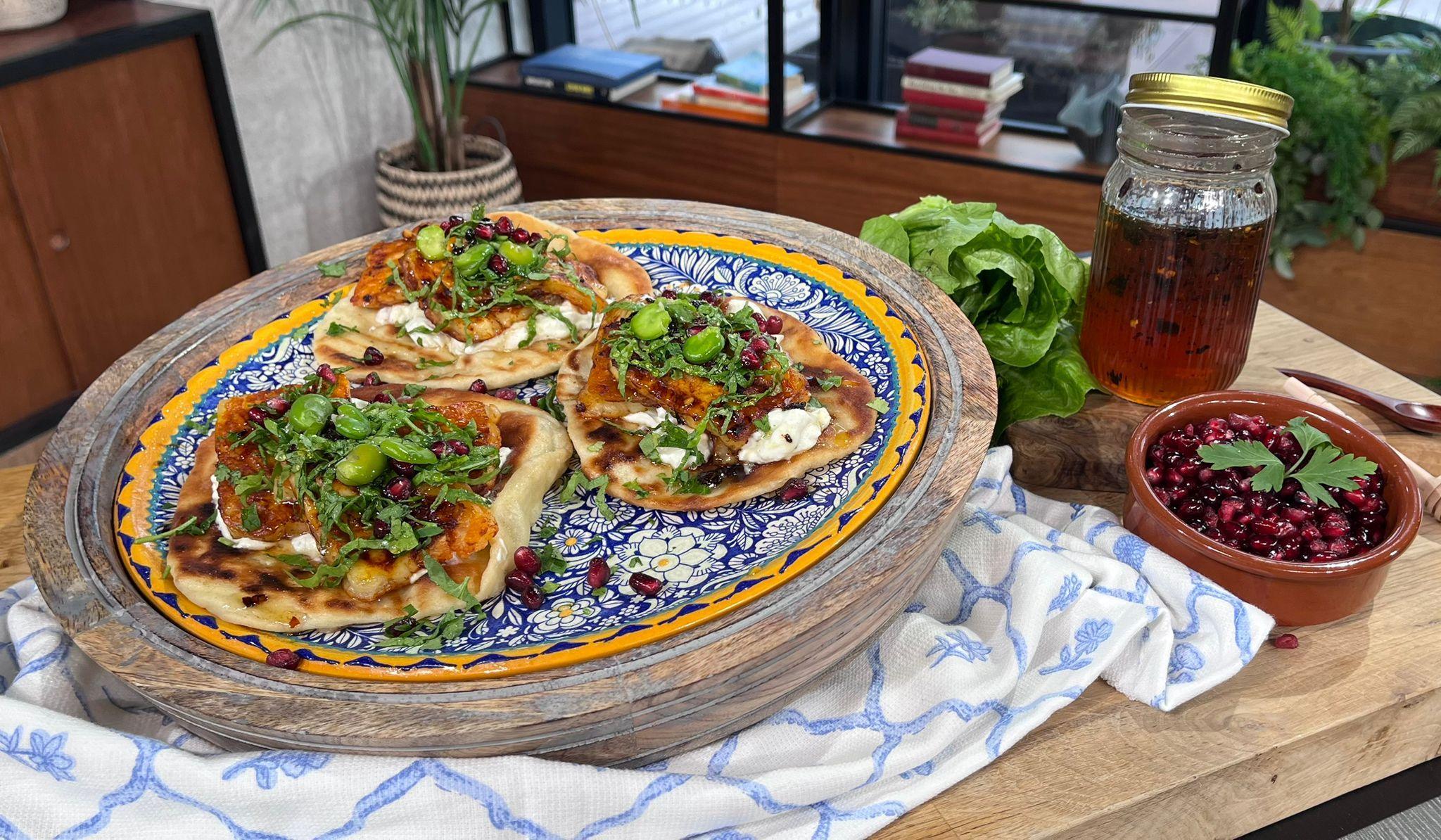 Three flatbreads on a raised plate, topped with halloumi, pomegranate seeds. To the left there is hot honey in a jar and a bowl of pomegranate seeds.