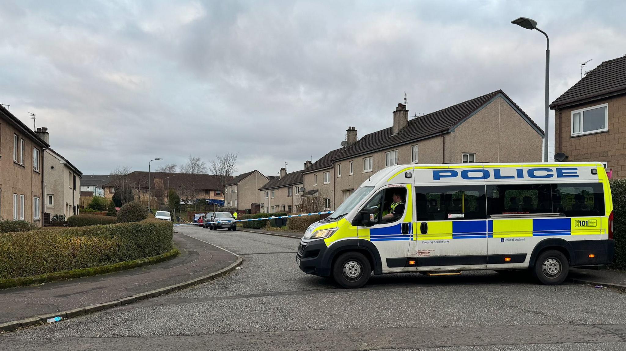 A police van blocks off a residential road. Police tape can be seen stretching across the road. The houses are covered in grey render. 