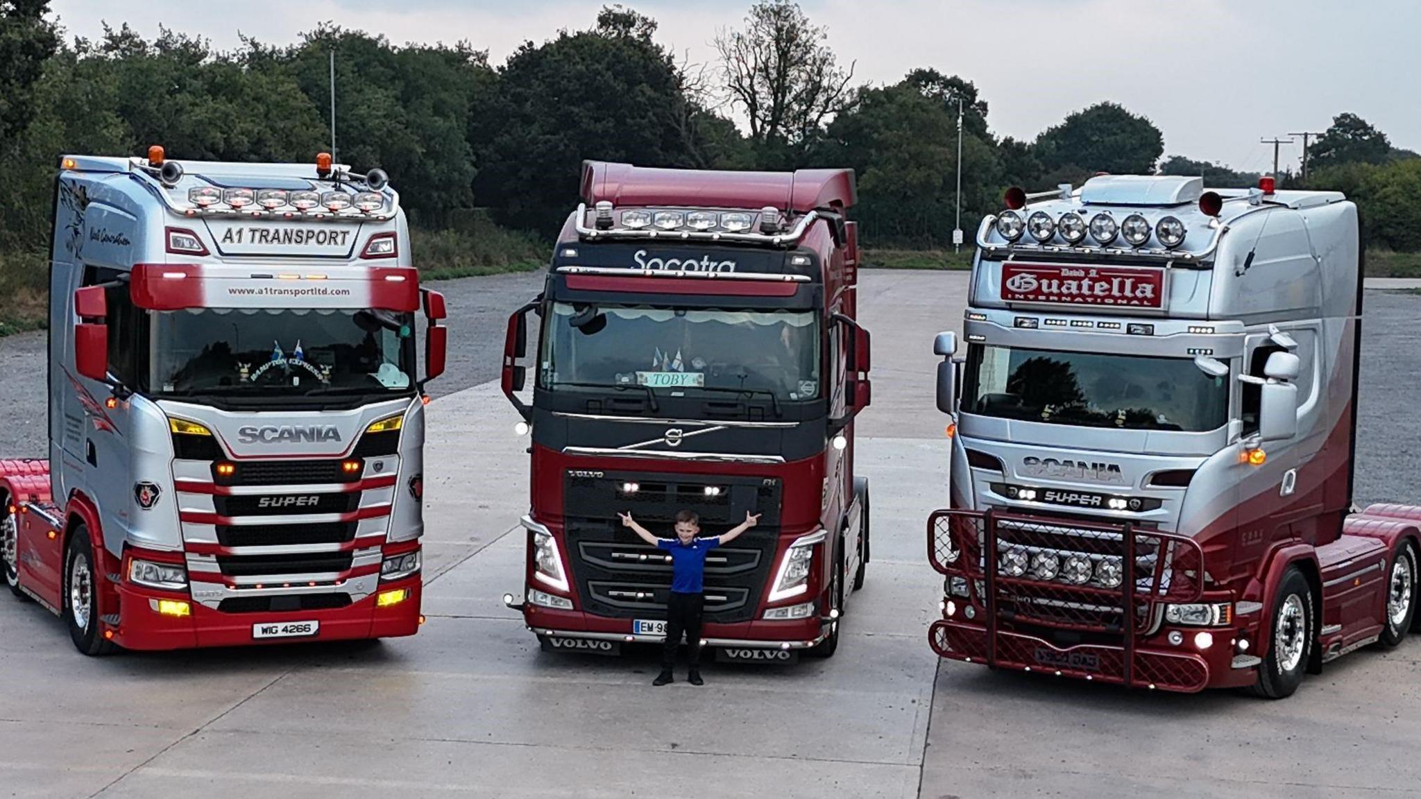 A boy in a royal blue t-shirt and dark trousers standing in front of the middle one of three trucks, with his arms spread wide. The red, maroon and silver trucks, which are not carrying loads, are standing on a wide forecourt with trees in the background.