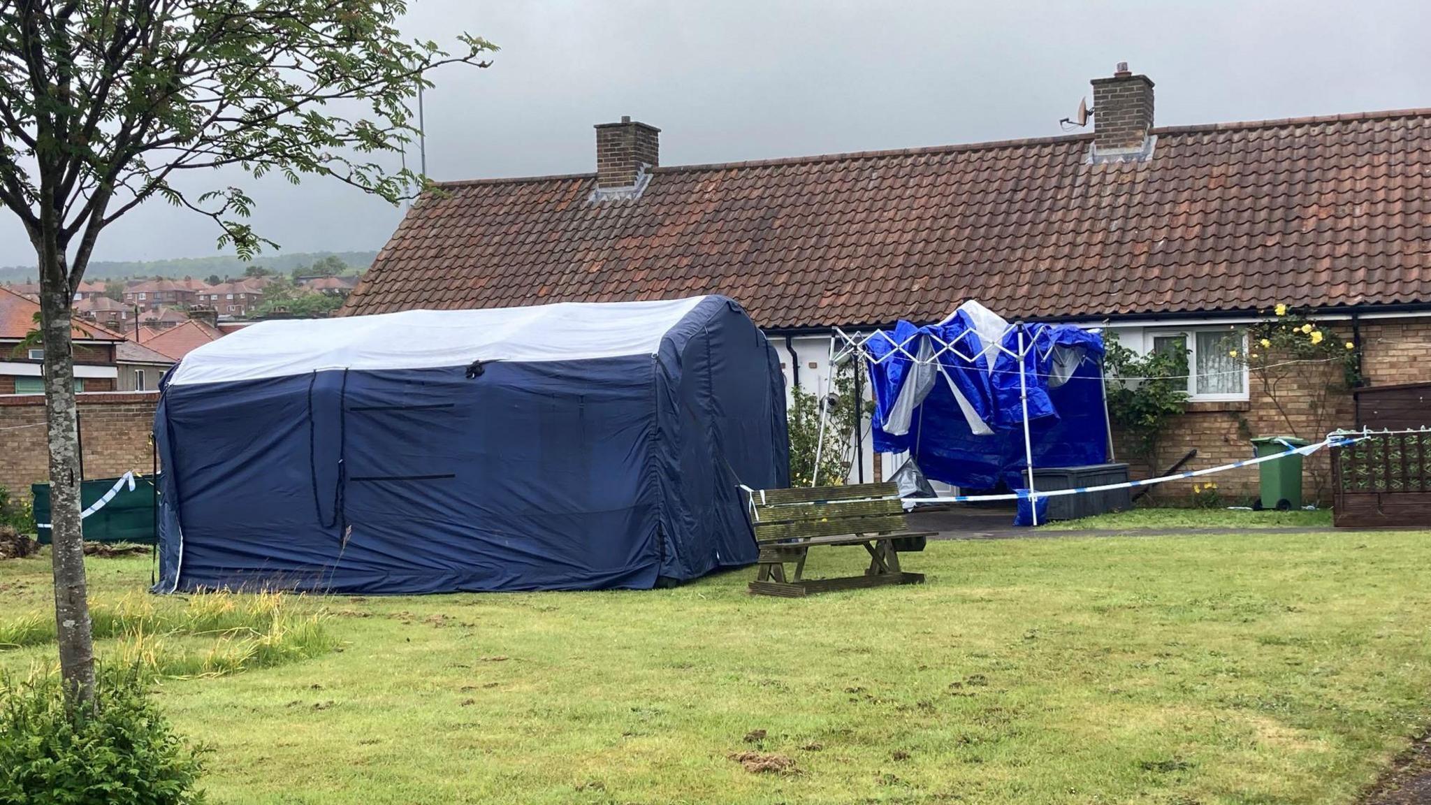 Two blue police tents and a cordon are set up outside bungalows.