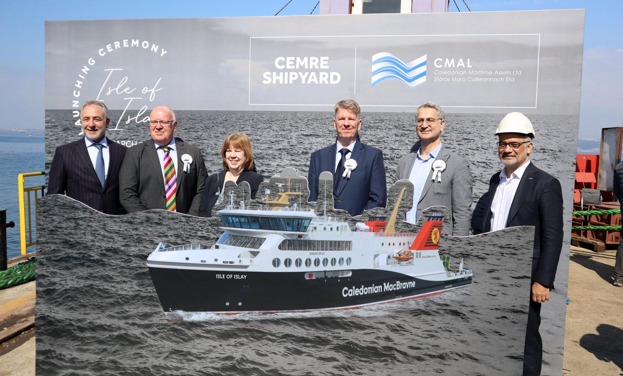 A group of five men in suits and woman standing behind a cardboard cutout of the new CalMac ferry MV Isle of Islay.