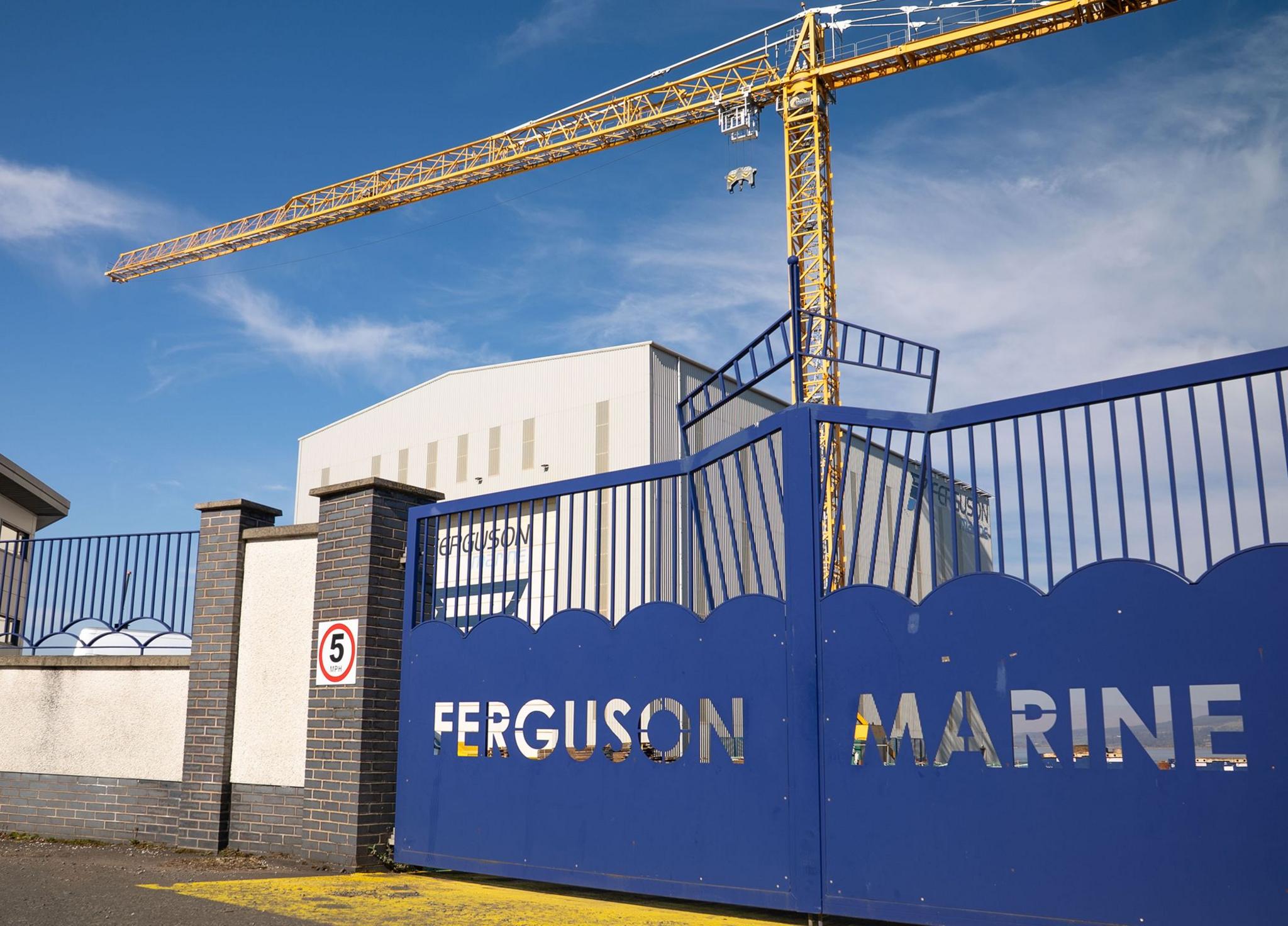 A blue gate with the words Ferguson Marine cut out. A large module hall and yellow crane can bee seen in the background