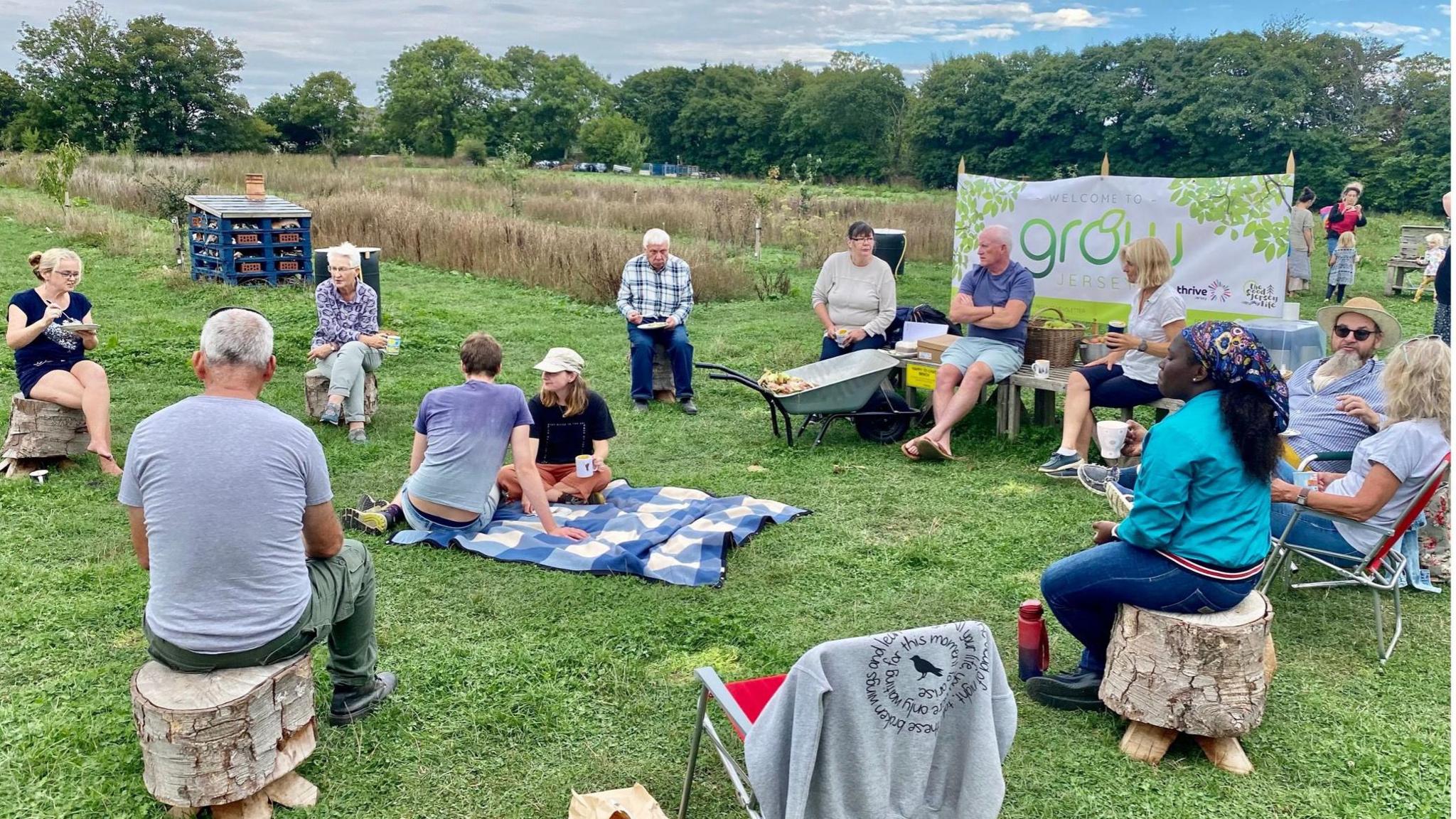 People sat on logs and chairs in a field in a circle. They are in a grass field.